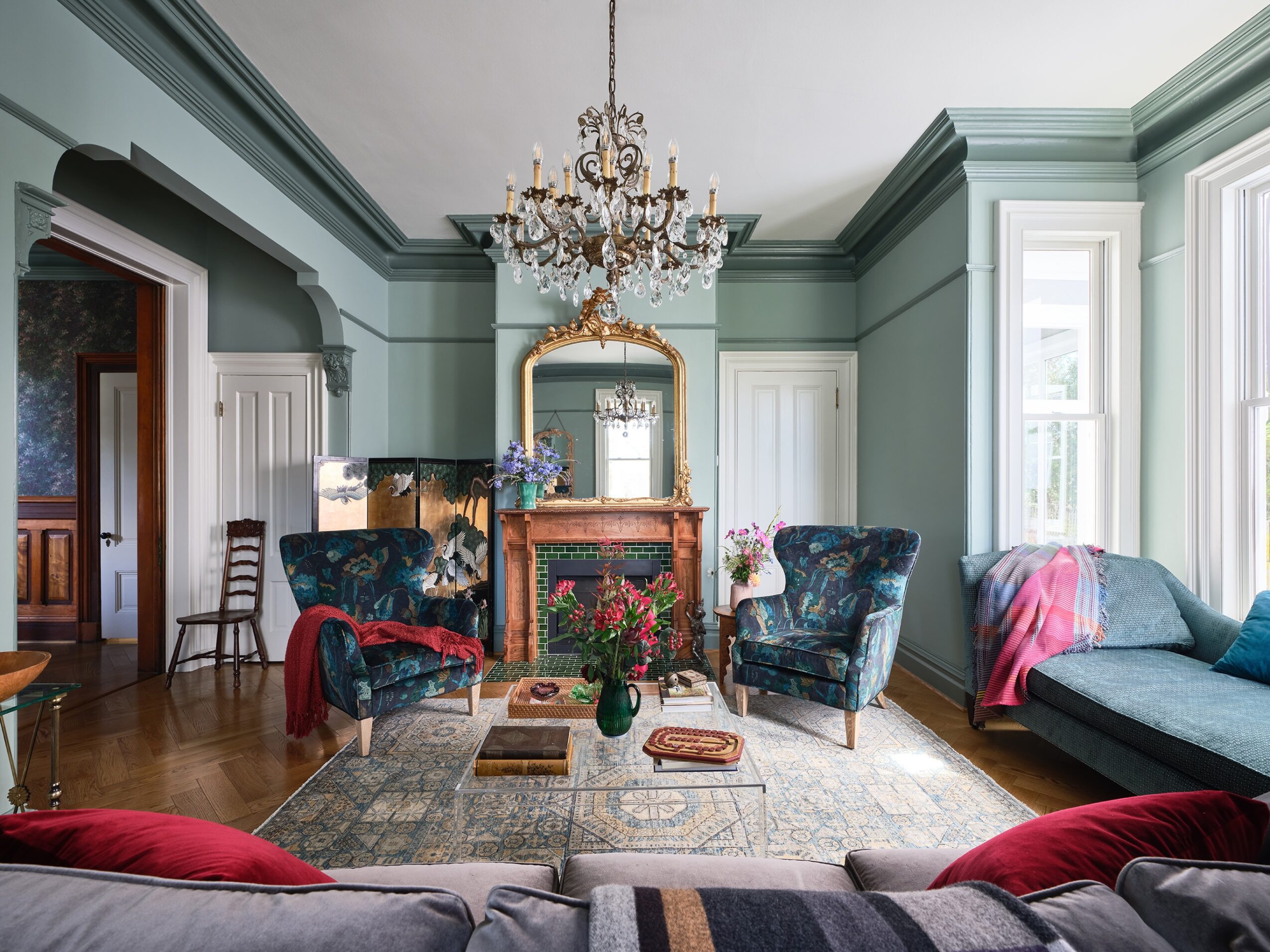 Elegant living room with a chandelier, fireplace, two armchairs, a sofa, and large windows, decorated in pastel green and blue tones.