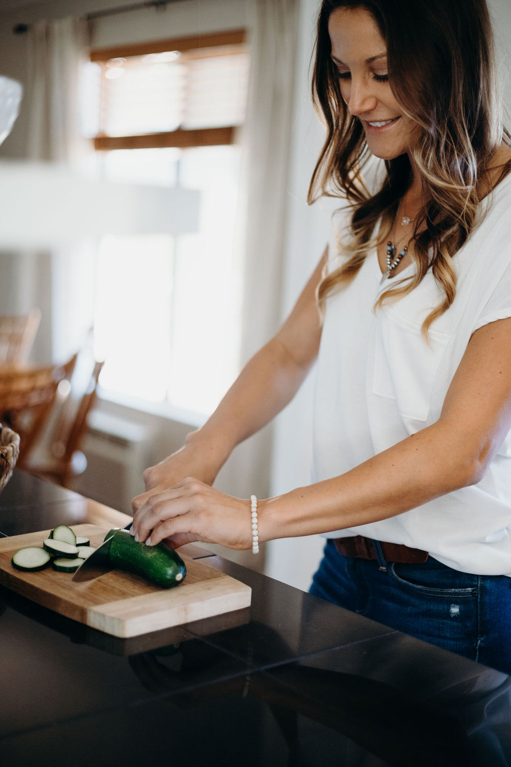 Woman smiling while preparing food in a bright kitchen, cutting vegetables on a wooden board, wearing a white shirt.
