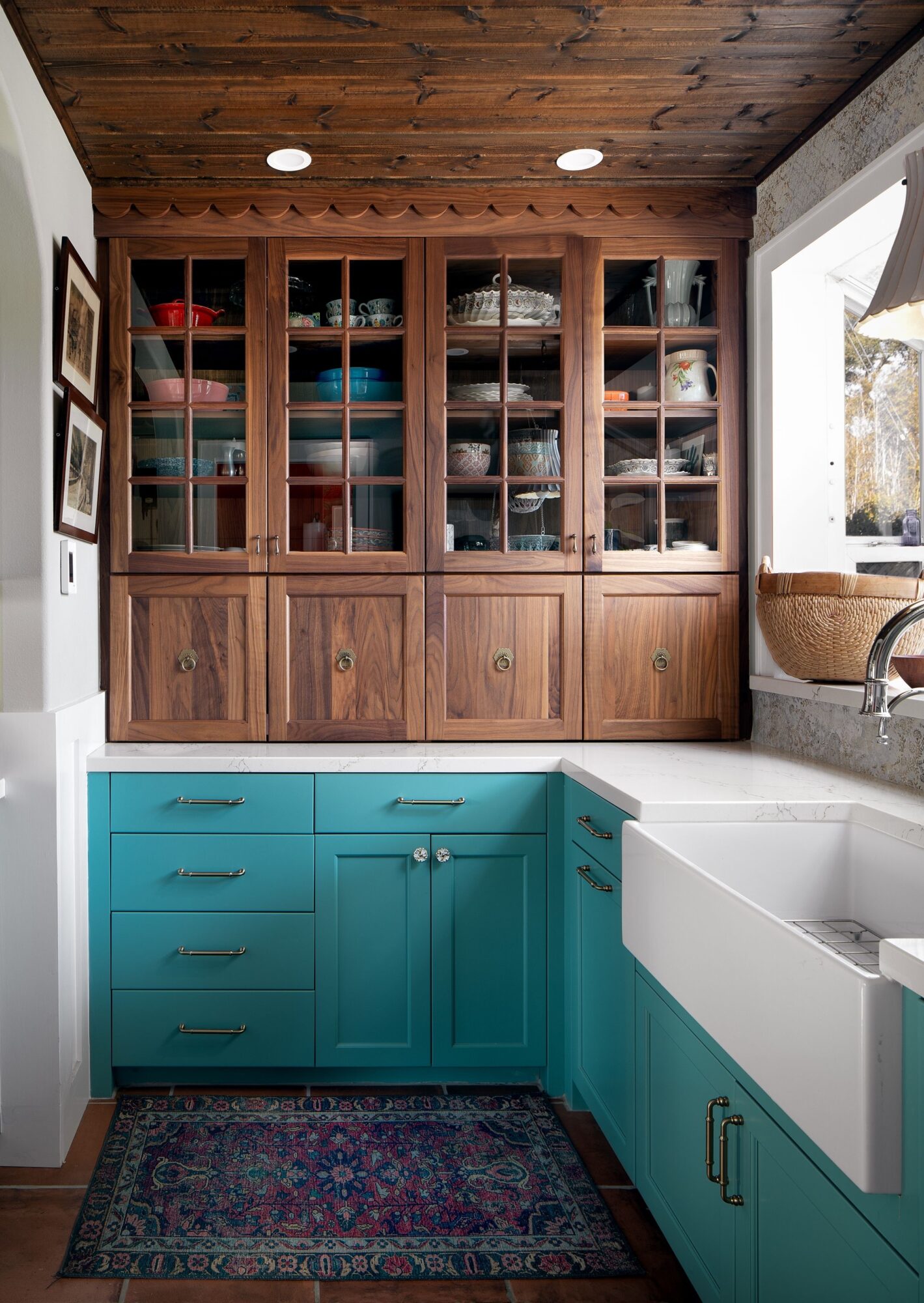 Kitchen with wooden upper cabinet, turquoise lower cabinets, white sink, window, and patterned rug.