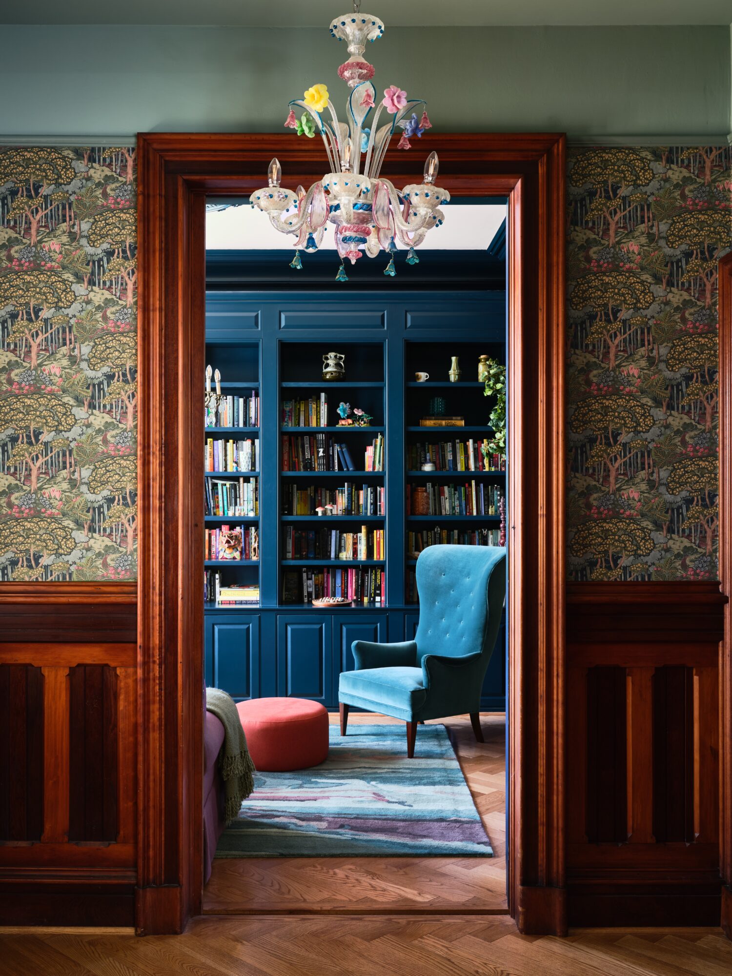 View through doorway into a room with blue bookshelves, a blue armchair, and a chandelier, with patterned wallpaper and wooden trim.