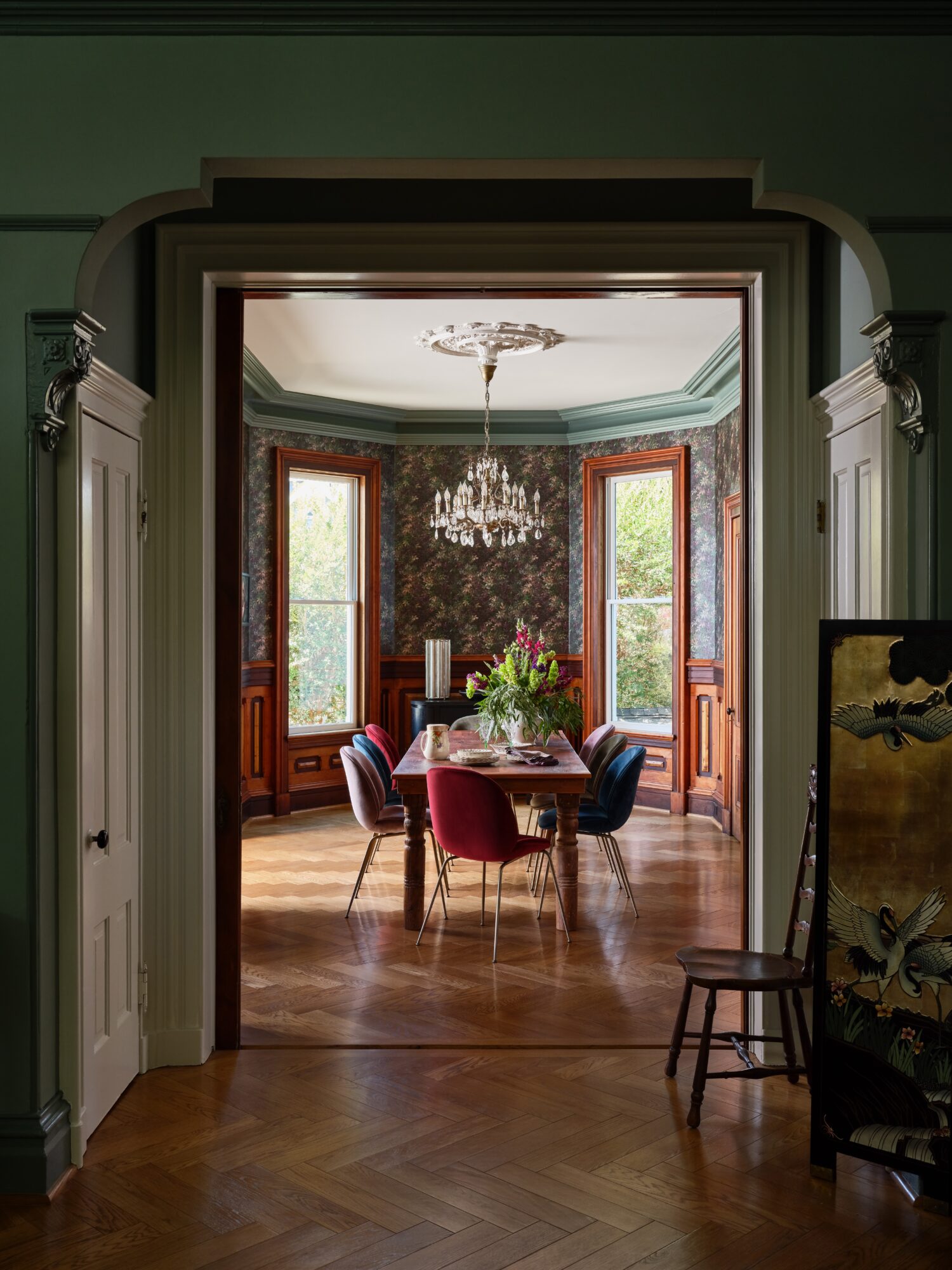 View through doorway into a dining room with a chandelier, table, chairs, and large windows, with a painting on the right.