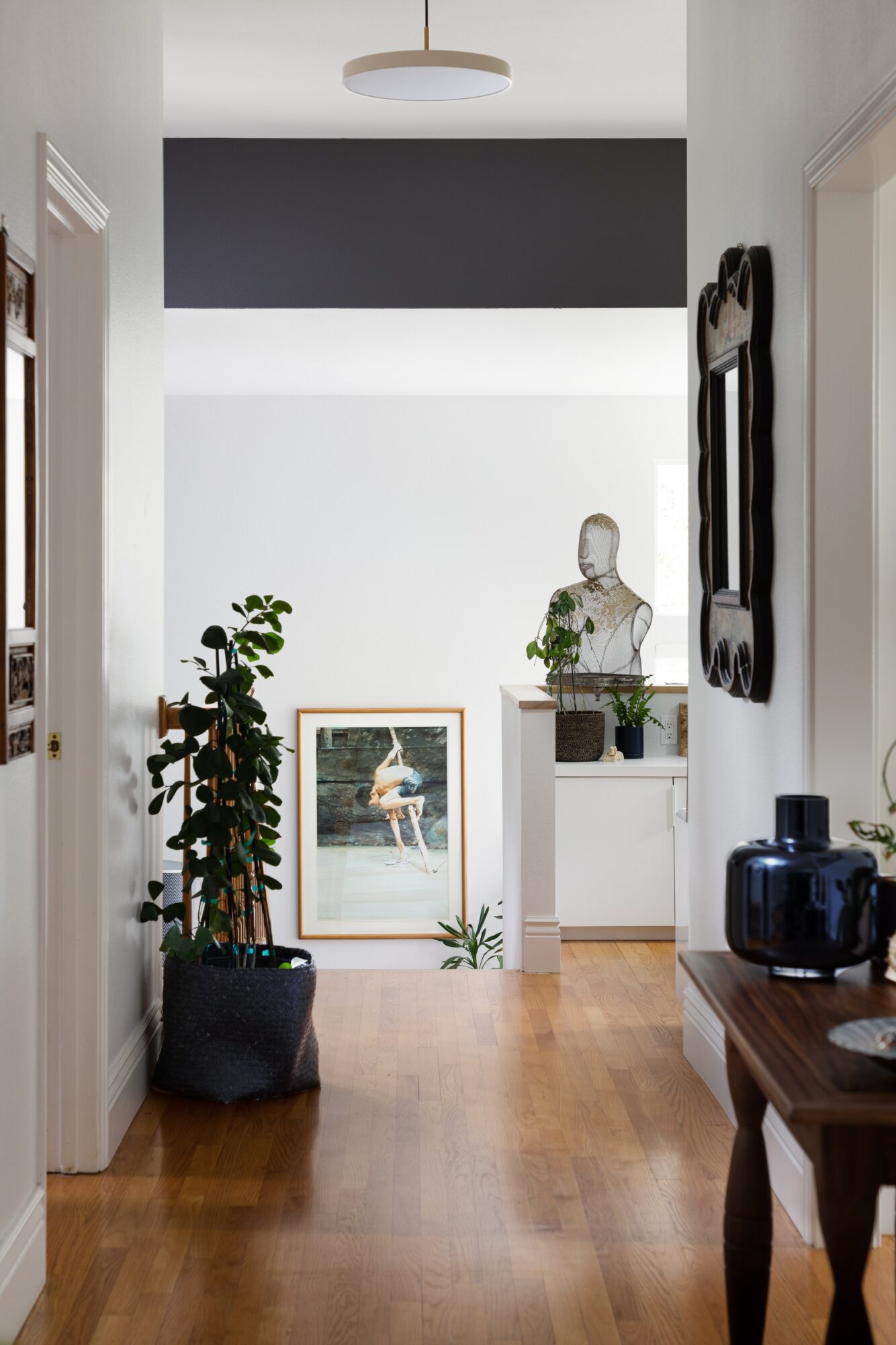 Hallway with wooden floor, potted plant, framed artwork, and decorative bust on a pedestal, with a mirror on the wall.