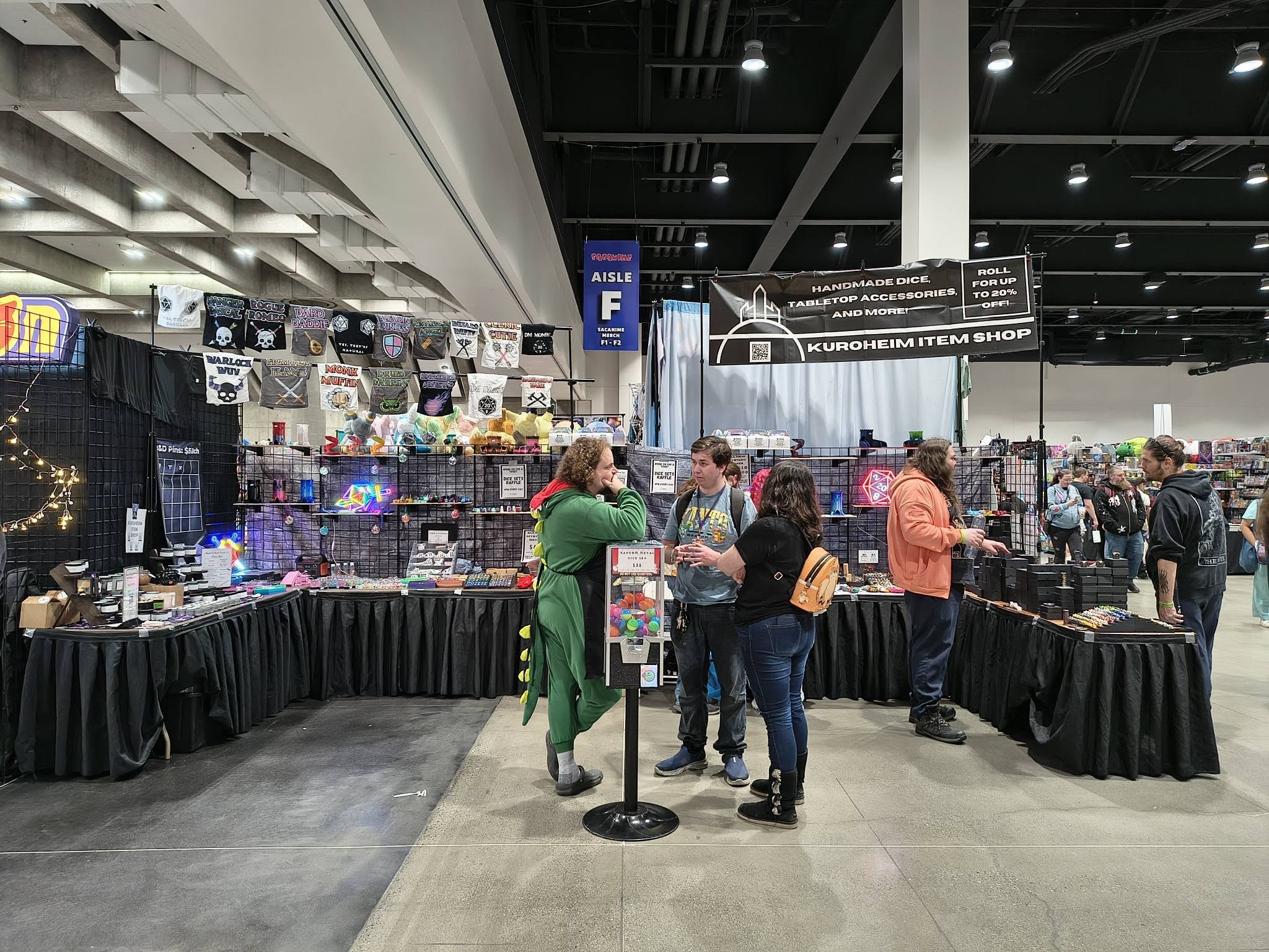 People browsing a booth with merchandise at an indoor event, with tables displaying items and signs overhead.
