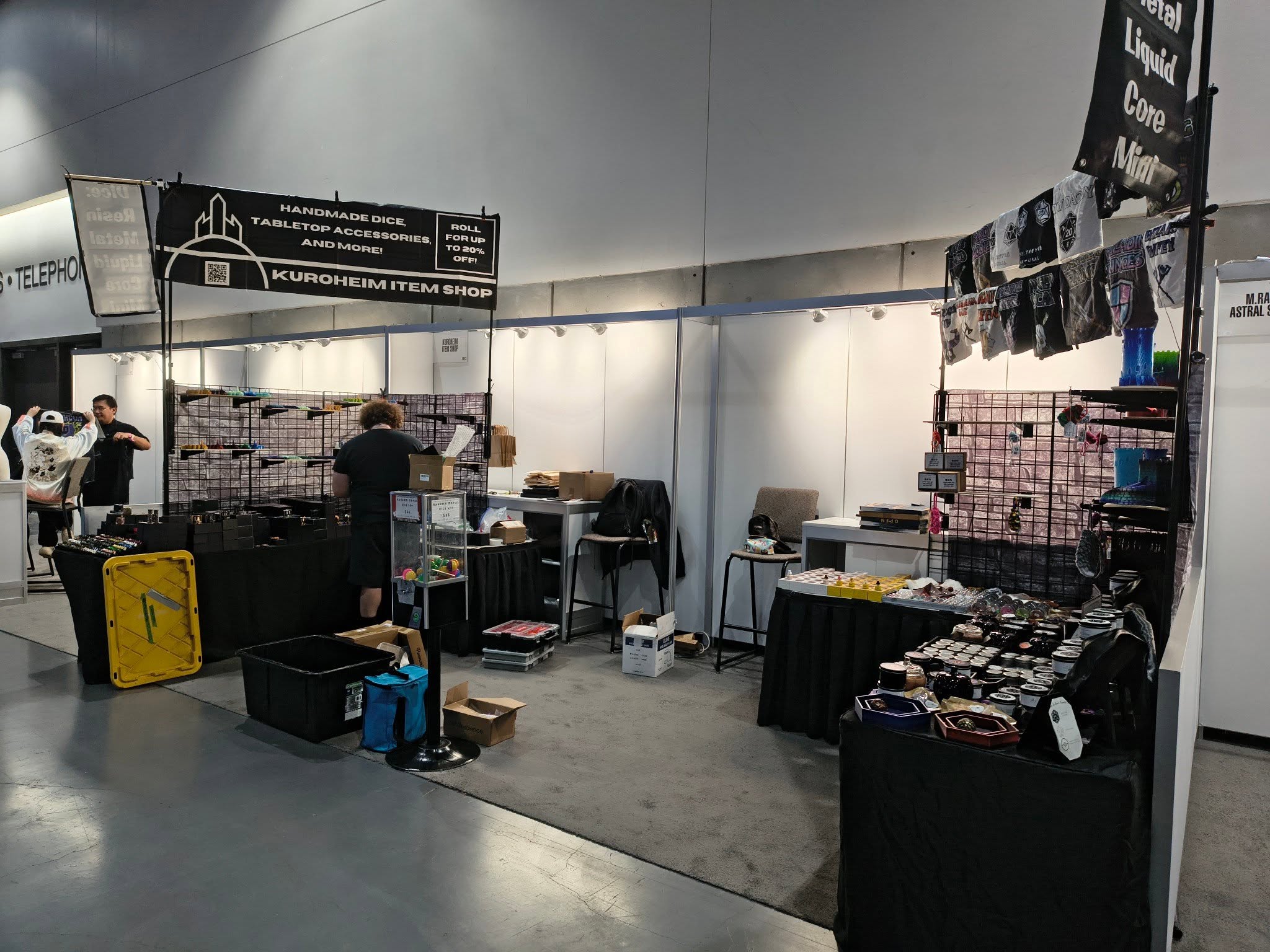 Vendor booth with various items displayed, people browsing, and a gray wall background.