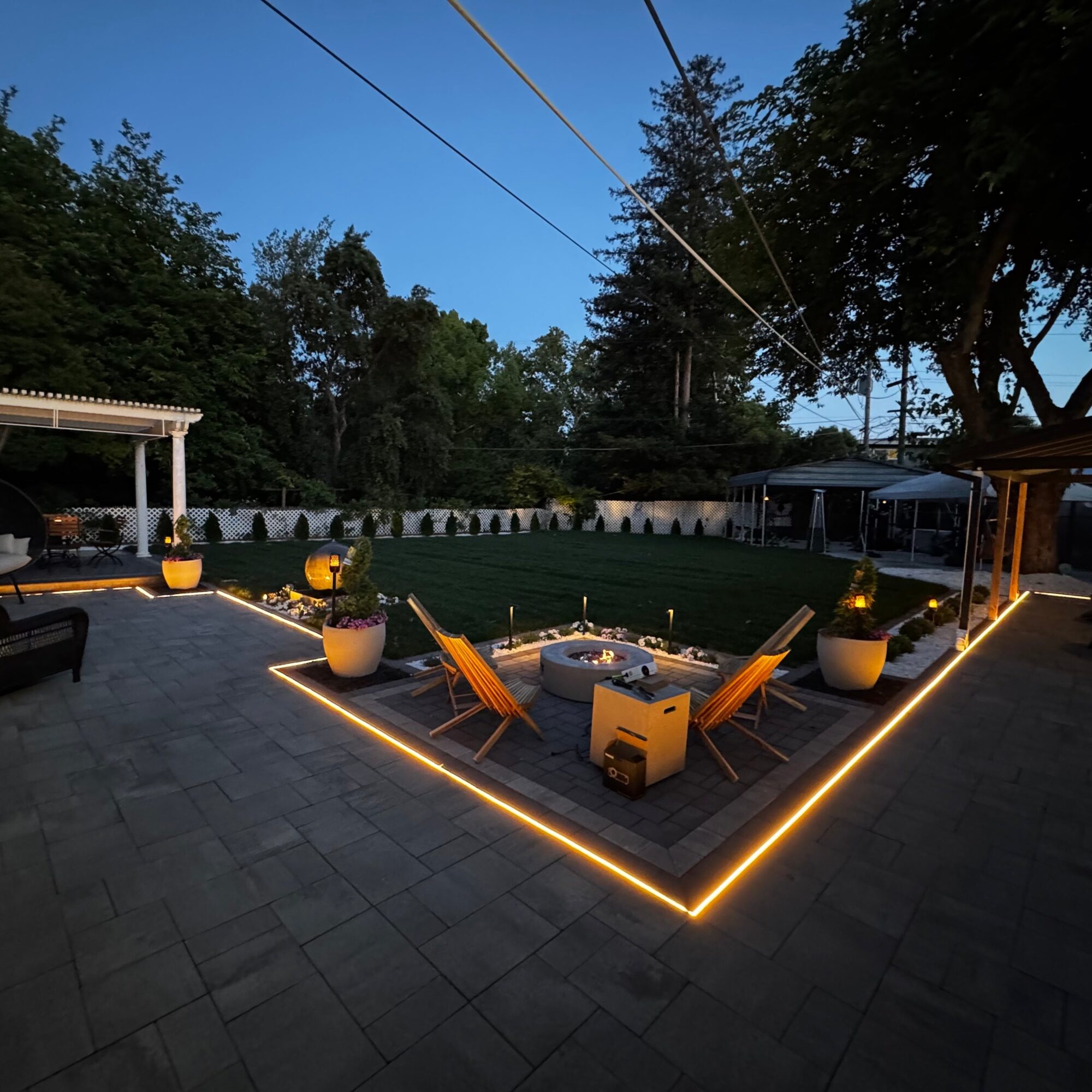 Outdoor patio with chairs, fire pit, and string lights, surrounded by trees and a lawn, at dusk.