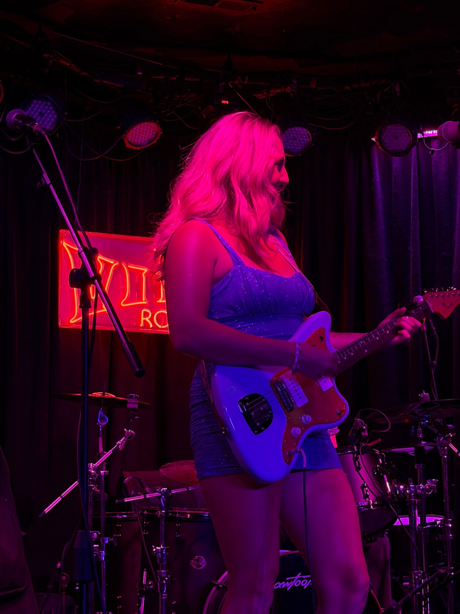 Person with long hair playing an electric guitar on stage with purple lighting.
