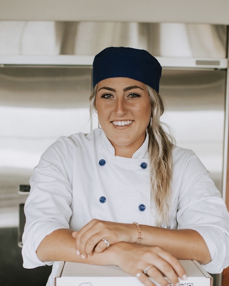 Smiling female chef in white uniform and navy hat, sitting at a kitchen counter with arms crossed.