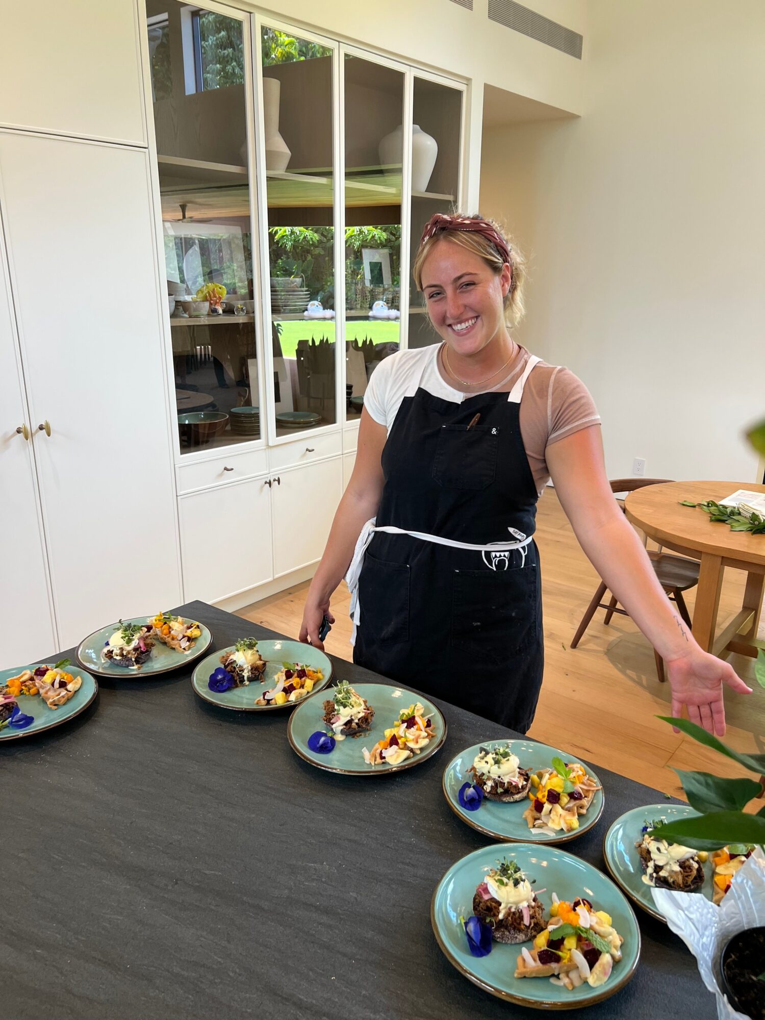 Young woman with short hair and glasses, wearing a black apron, standing in a kitchen with plates of decorated desserts.