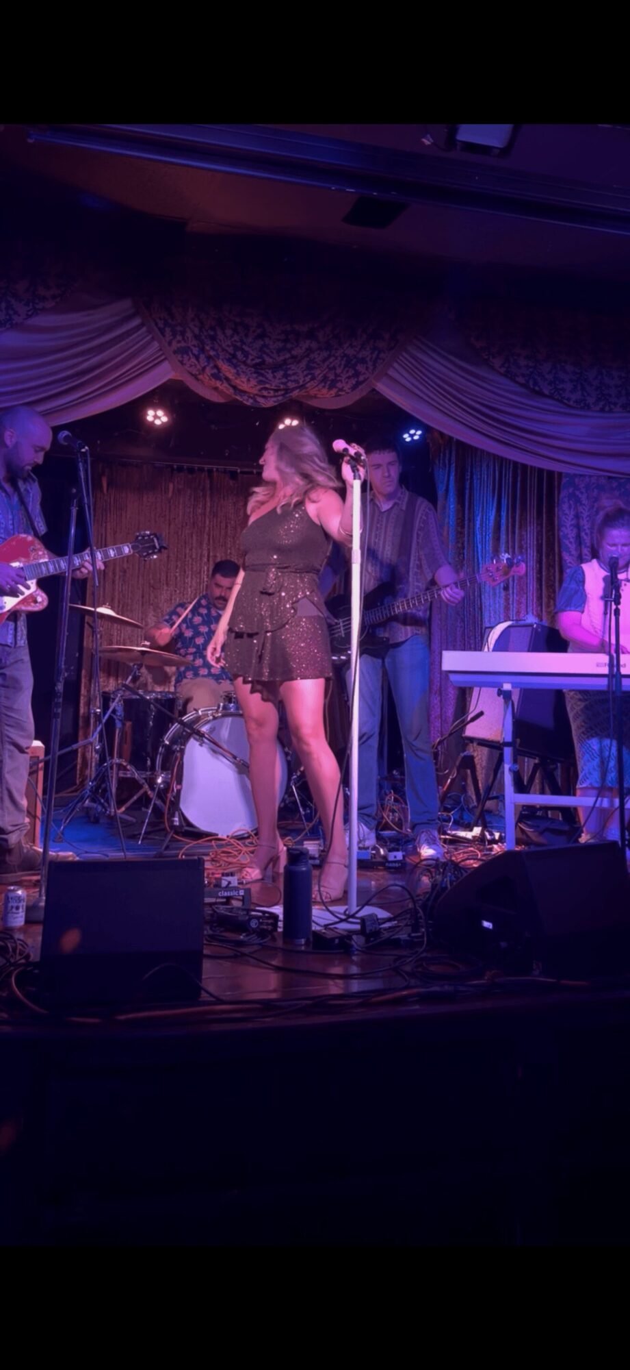 Musicians perform on stage with a singer, keyboard, drums, and guitar, under purple and blue stage lighting.