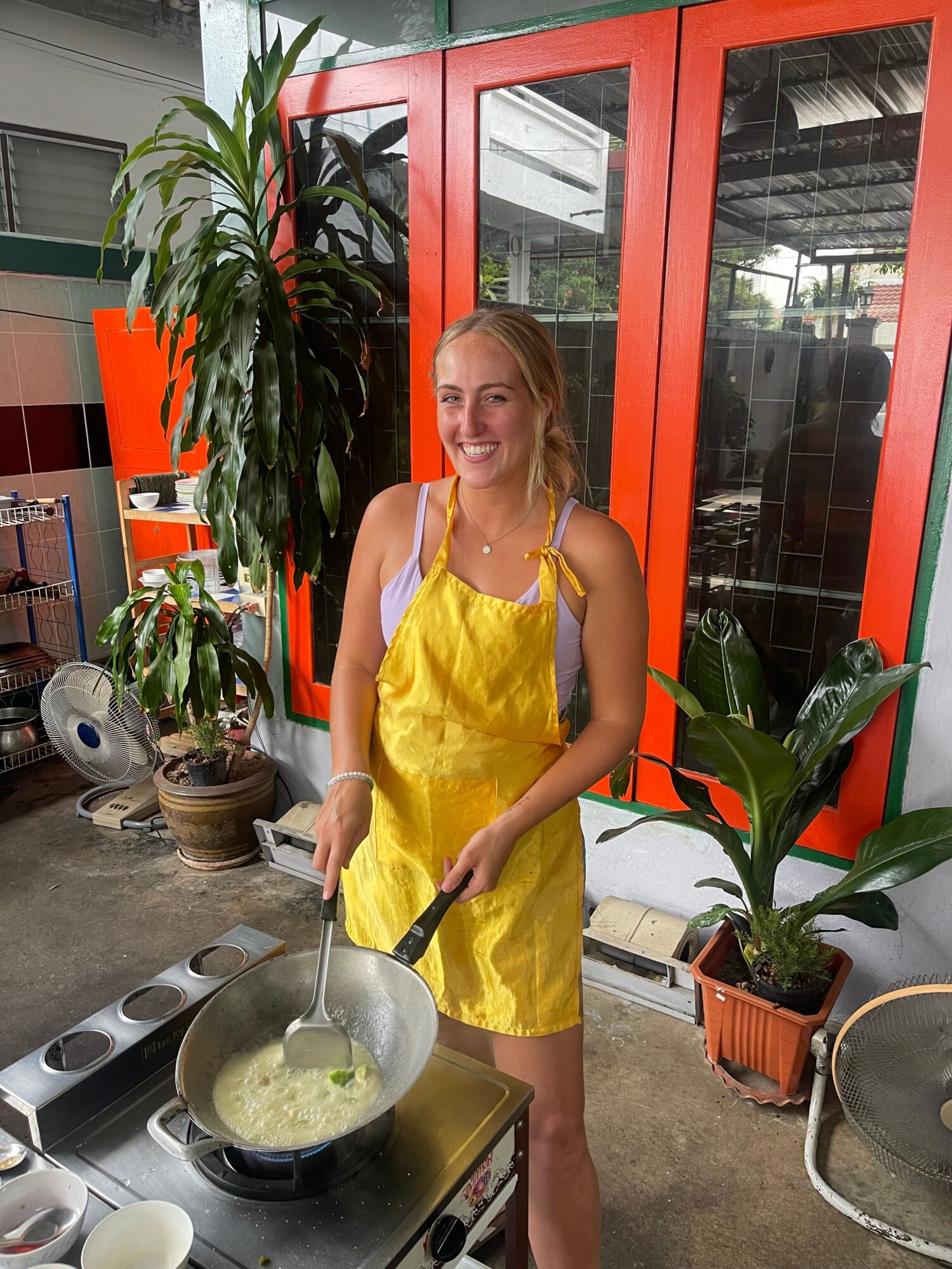 Woman in yellow dress cooking at outdoor stove with plants and a glass door behind her.