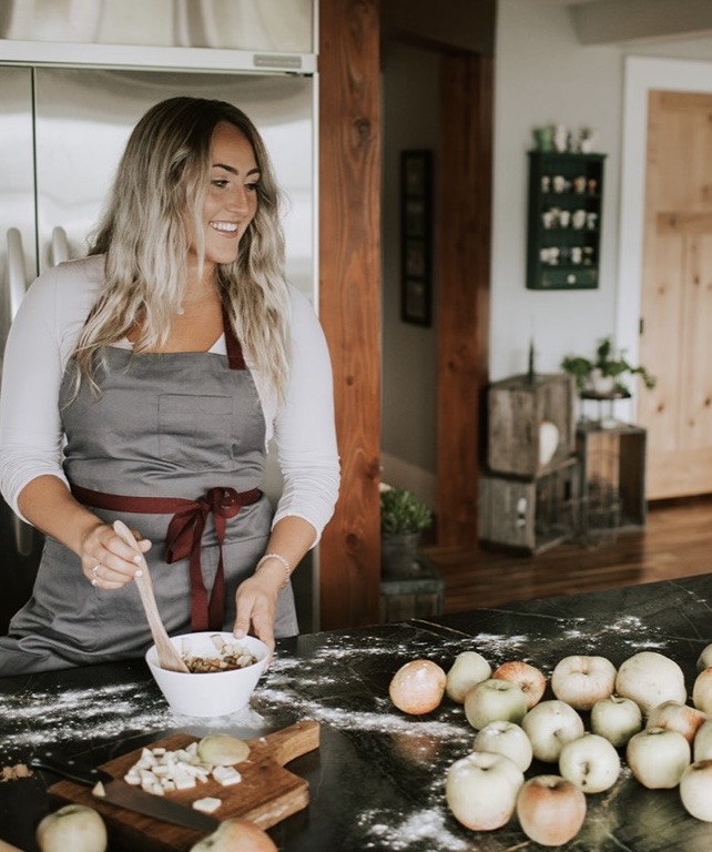 Woman with long hair wearing an apron, smiling, mixing ingredients in a bowl in a kitchen, with onions on the counter.