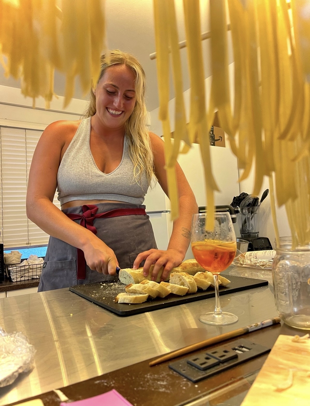 Woman slicing lemons in a kitchen with hanging pasta above, a glass of wine, and kitchen utensils nearby.