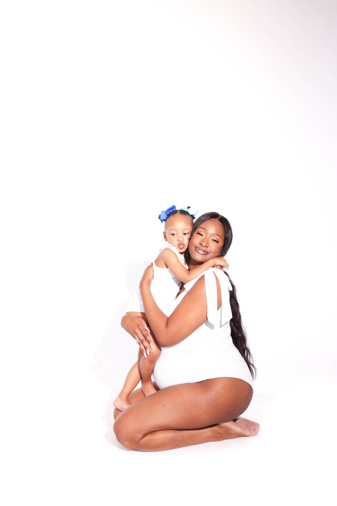 Woman sitting on the floor holding a smiling child with a blue bow in her hair, both wearing white outfits.