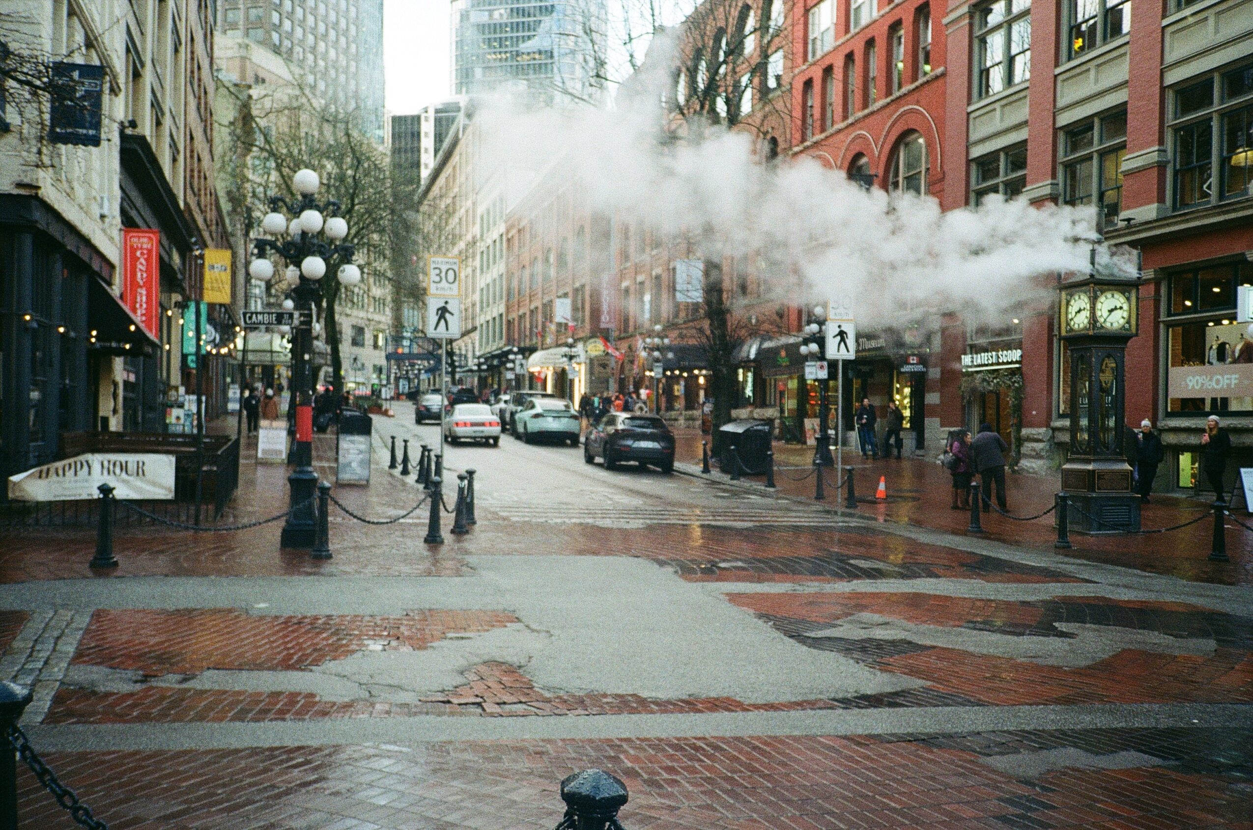 City street with buildings, cars, pedestrians, and steam rising from underground, wet pavement, and overcast sky.