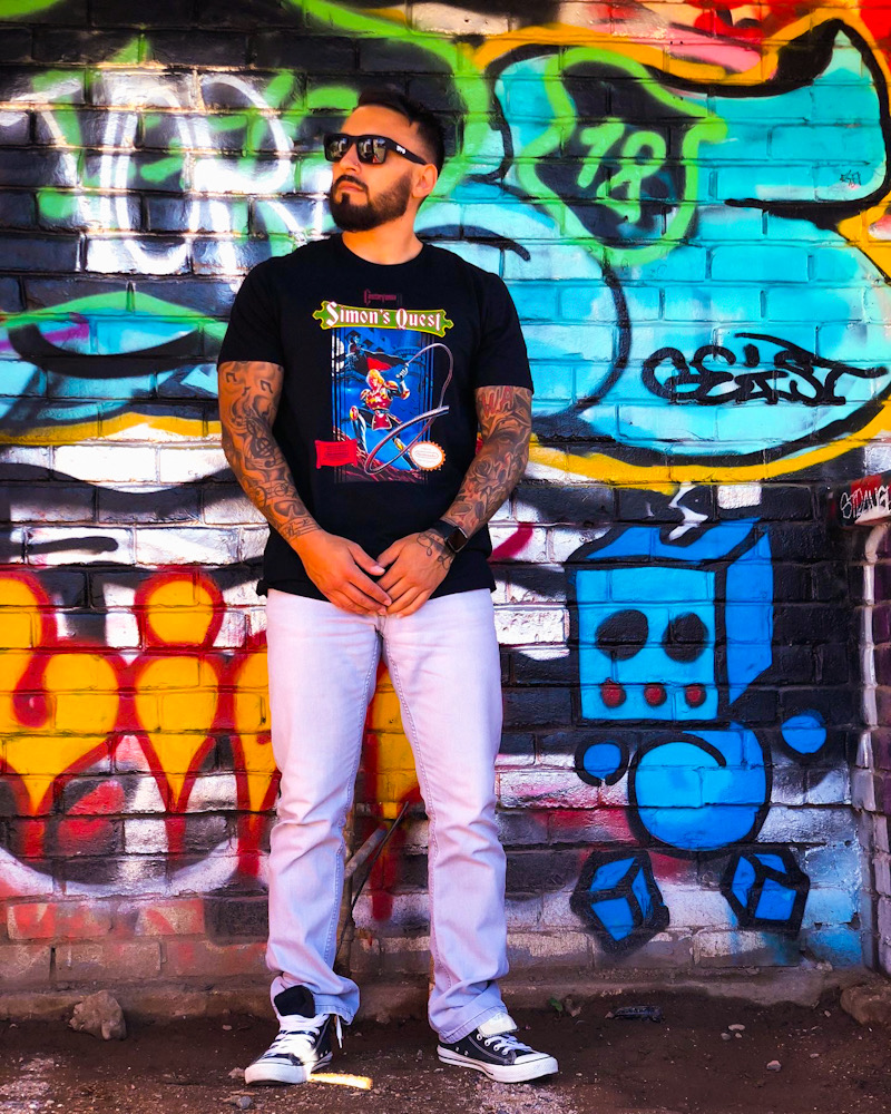 Man with sunglasses and tattoos stands in front of colorful graffiti wall, wearing a black graphic t-shirt and white pants.