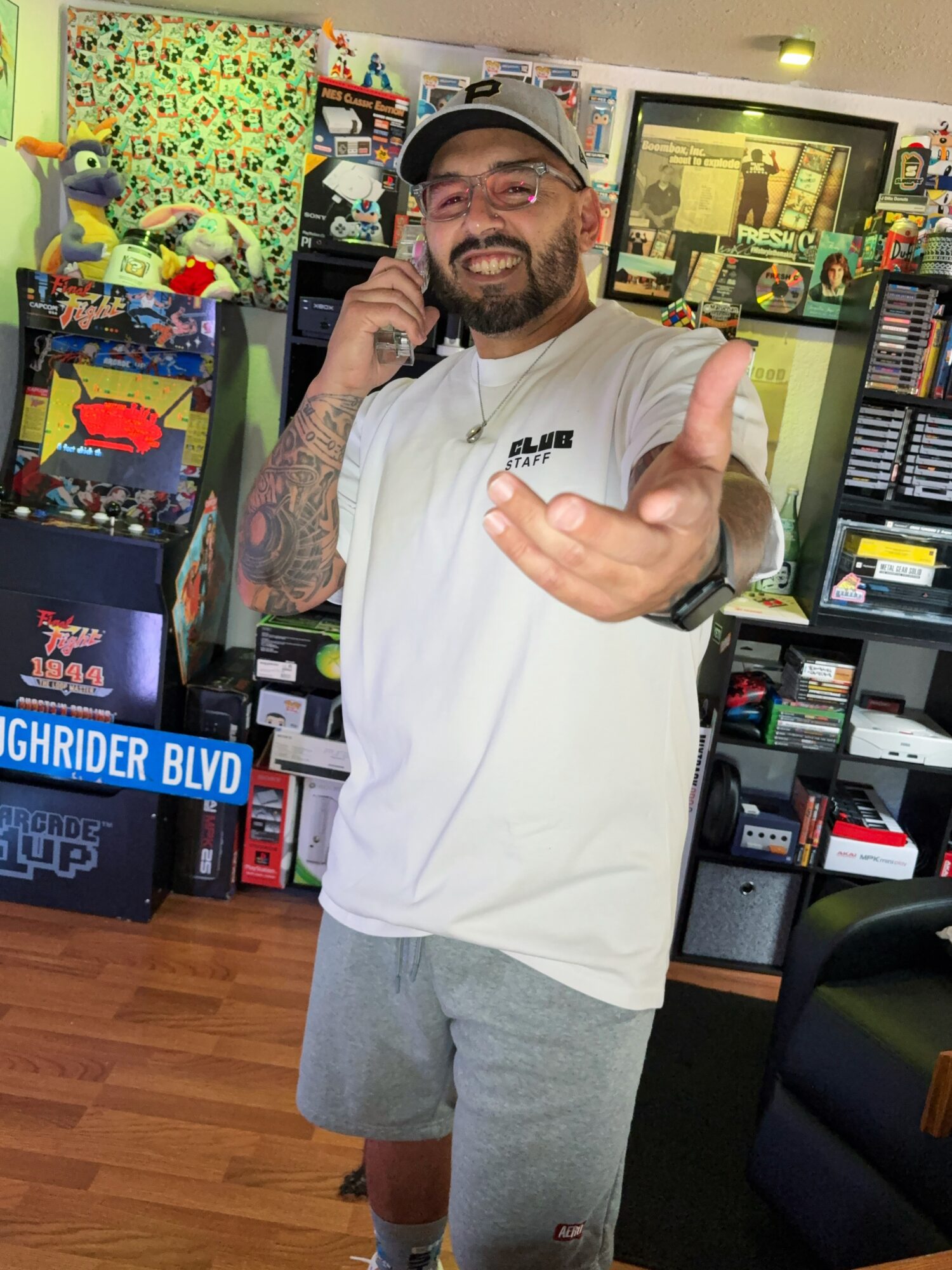 Smiling man with a beard, wearing a cap and white t-shirt, gesturing with his hand in a store filled with toys and electronics.