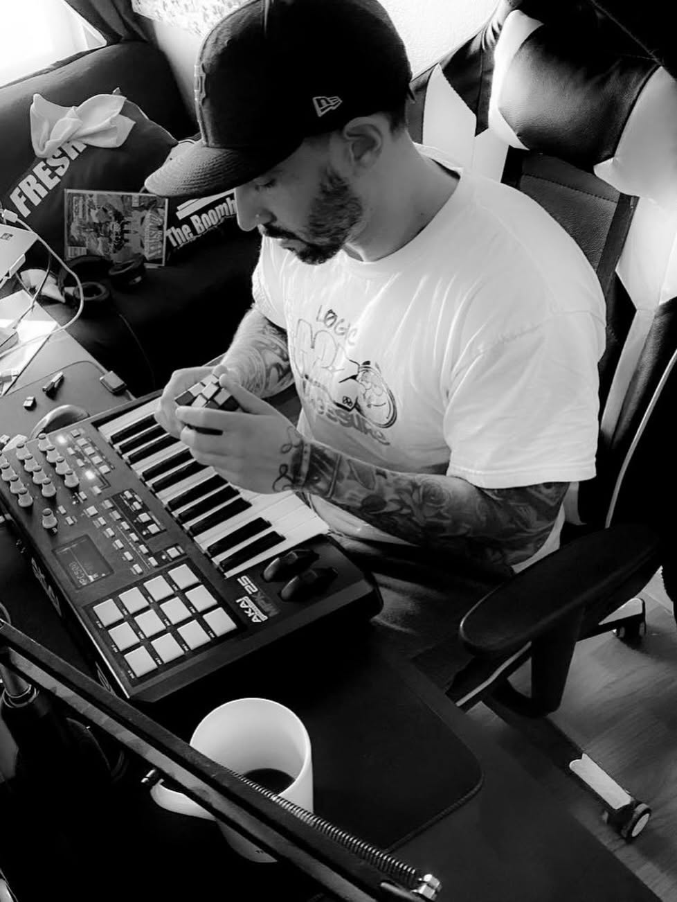 Man with tattoos wearing a cap and white T-shirt, sitting at a desk with music equipment, looking at his phone.