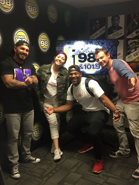 Four people smiling and posing in front of a radio station backdrop with a sign reading 'Power 98 & 101.9'.