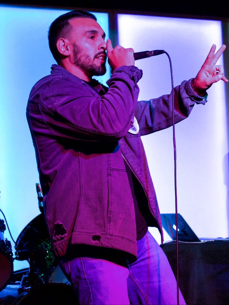 Person holding microphone, making peace sign with other hand, standing against illuminated background.