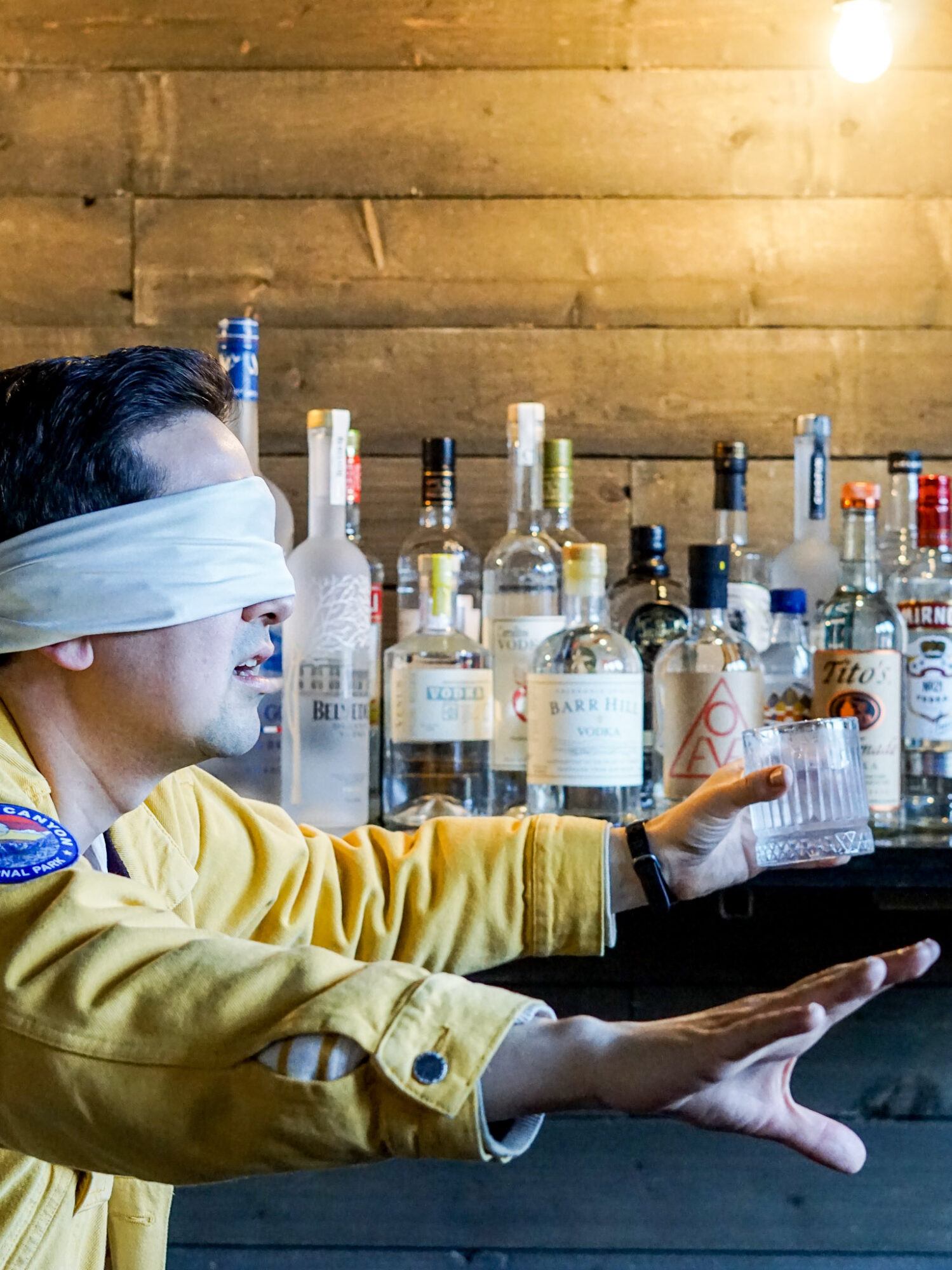 Person with a blindfold reaching for a glass at a bar with bottles in the background.