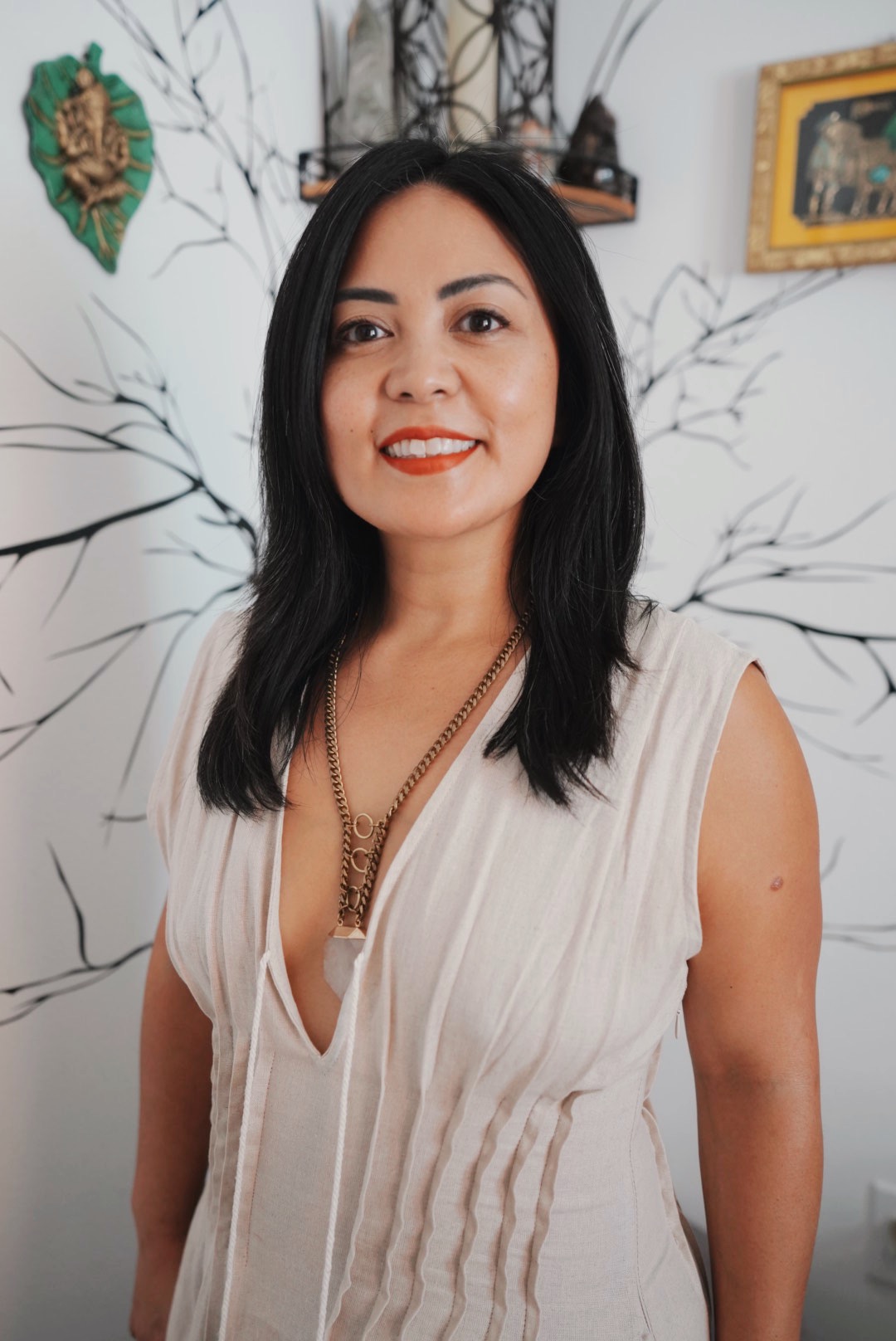Woman with black hair smiling, wearing a white sleeveless top, standing indoors with wall art and a shelf behind her.