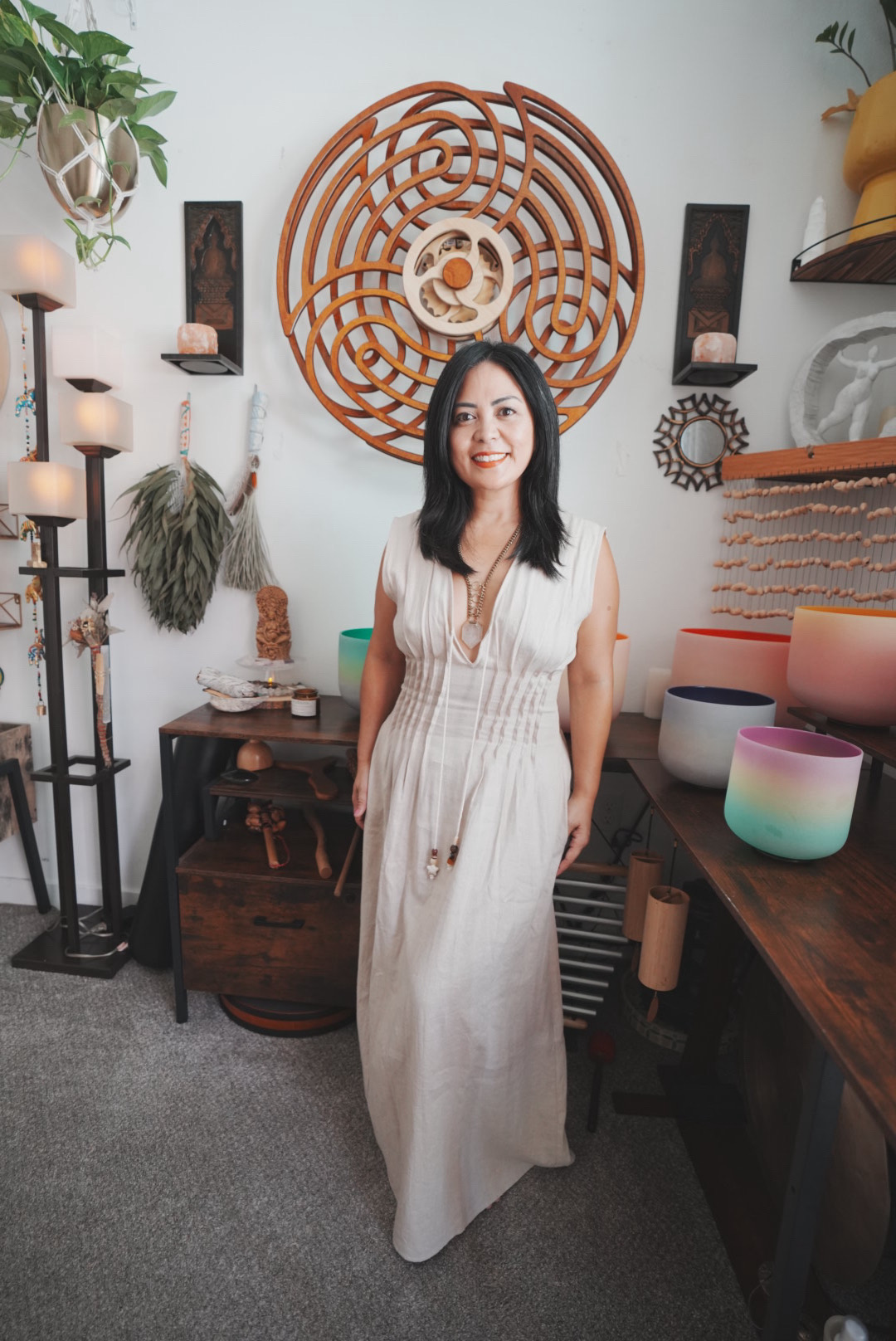 Woman in a white dress standing in a room with decorative wall art, plants, and shelves, smiling at the camera.
