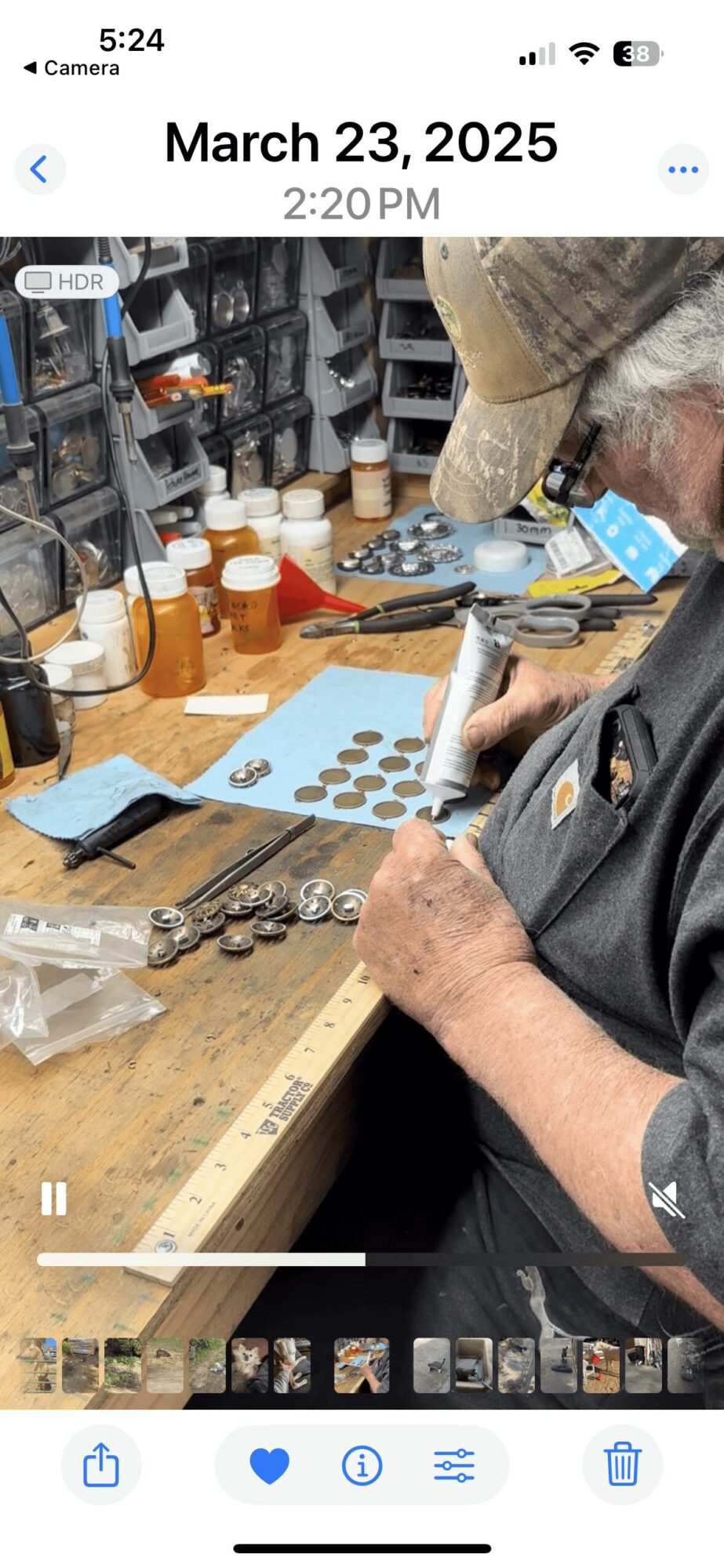 Person working at a cluttered workbench with small containers, tools, and jewelry components, wearing a hat and glasses.
