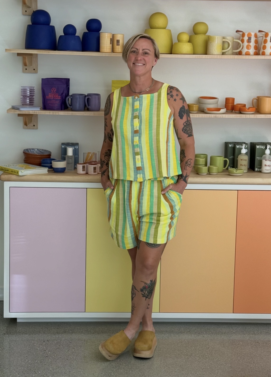 Woman with tattoos wearing a colorful striped outfit, smiling, standing in front of shelves with pottery and cups.