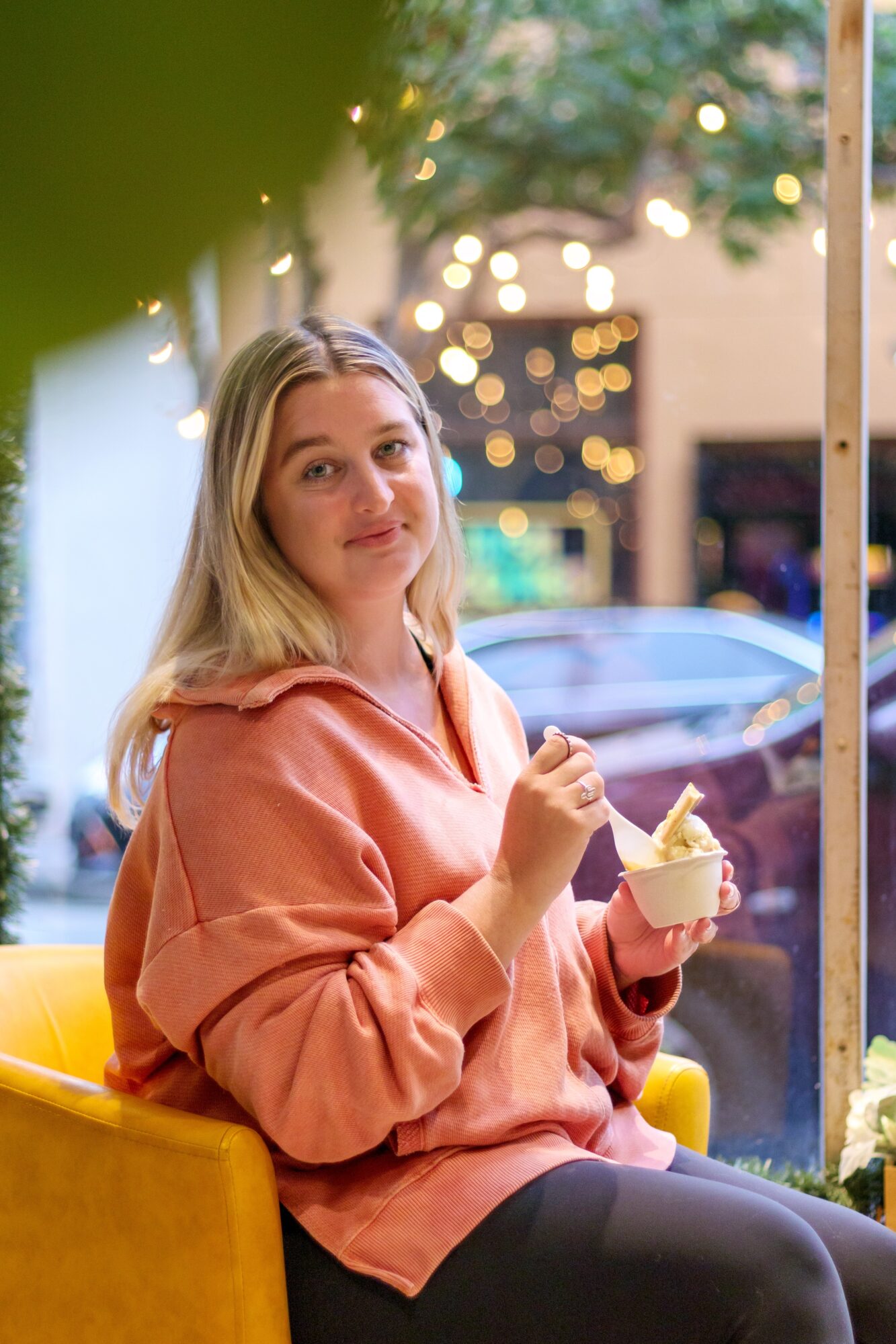 Young woman with long blonde hair in a pink hoodie sitting outdoors, holding a cup of ice cream, smiling.