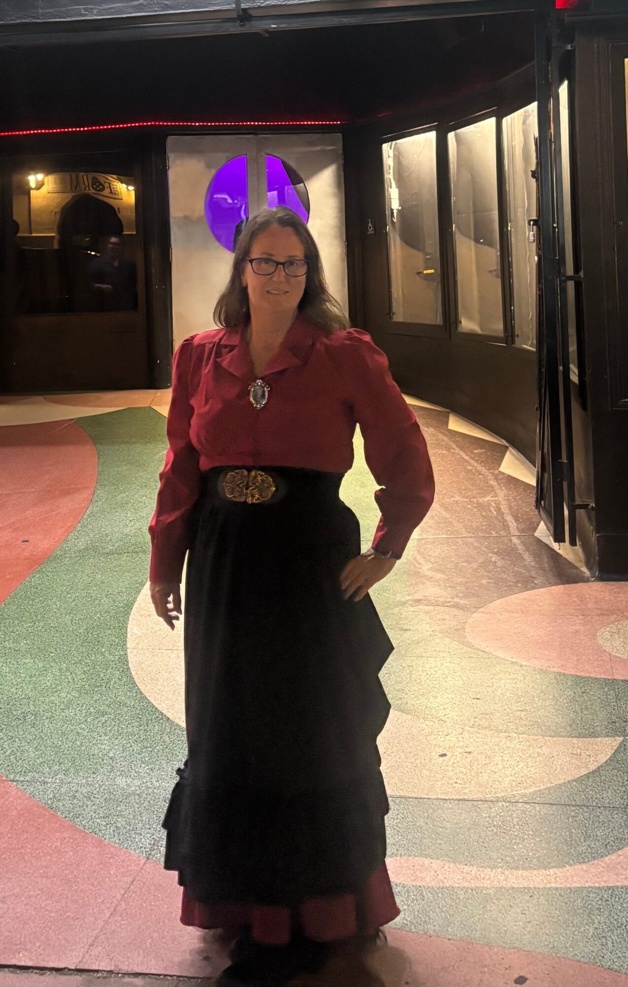 Woman in red blouse and black skirt standing indoors with colorful floor and purple illuminated sign behind her.