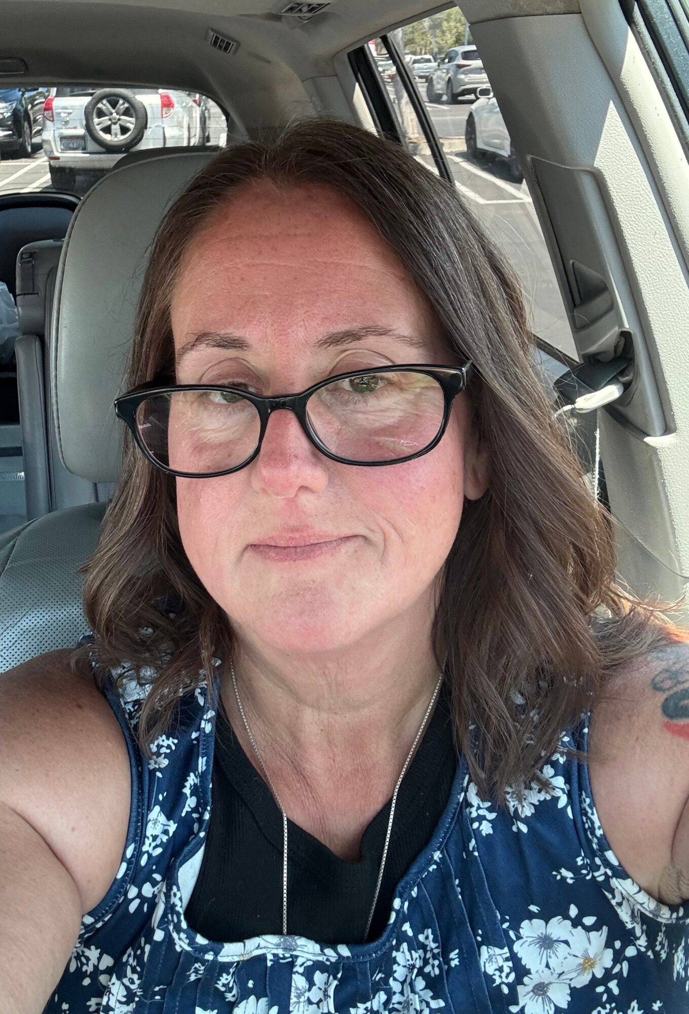 Woman with glasses and shoulder-length hair sitting in a car, wearing a blue floral top, looking at the camera.