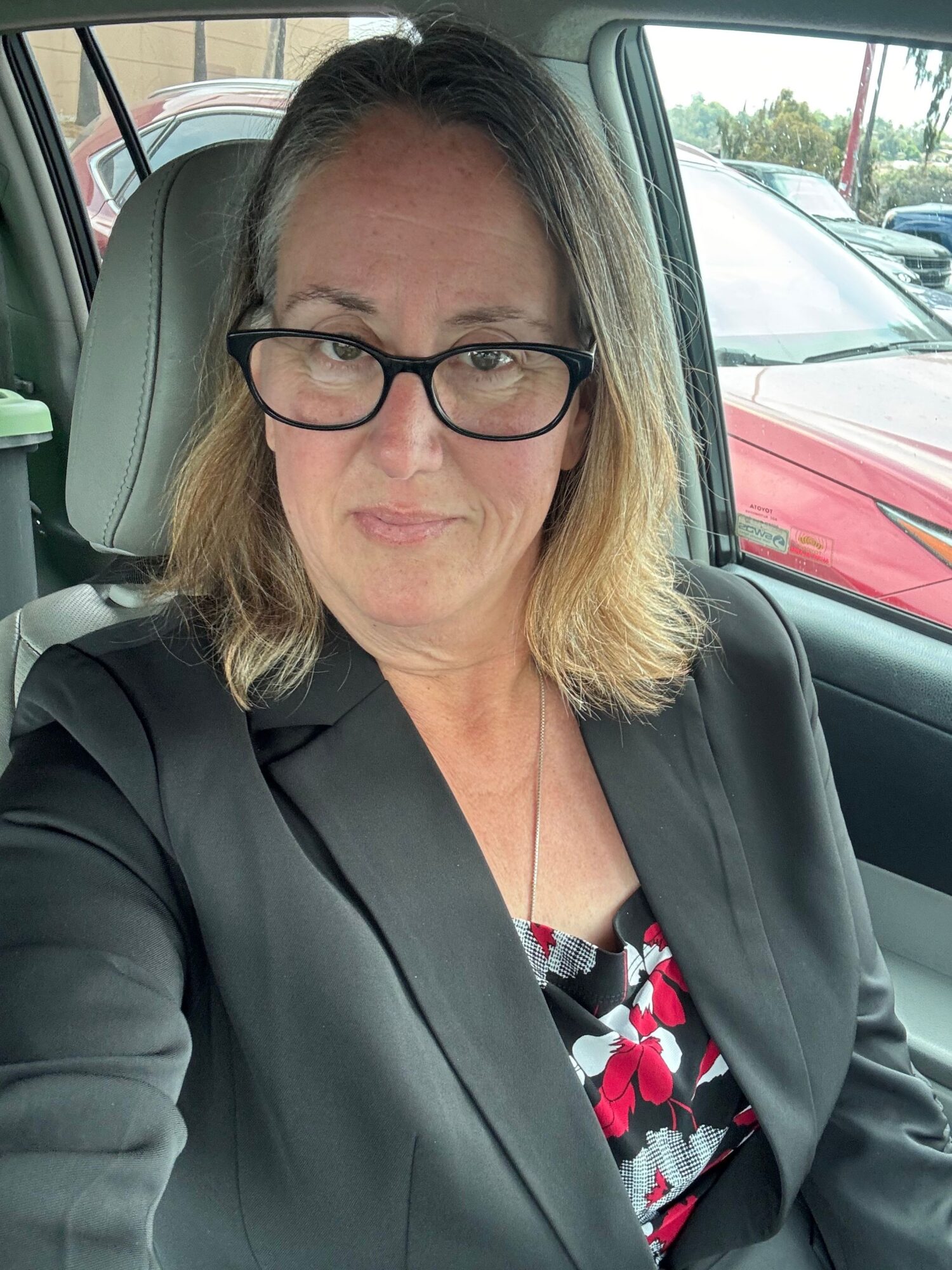 Woman with glasses sitting in a car, looking at the camera, with cars visible outside the window.