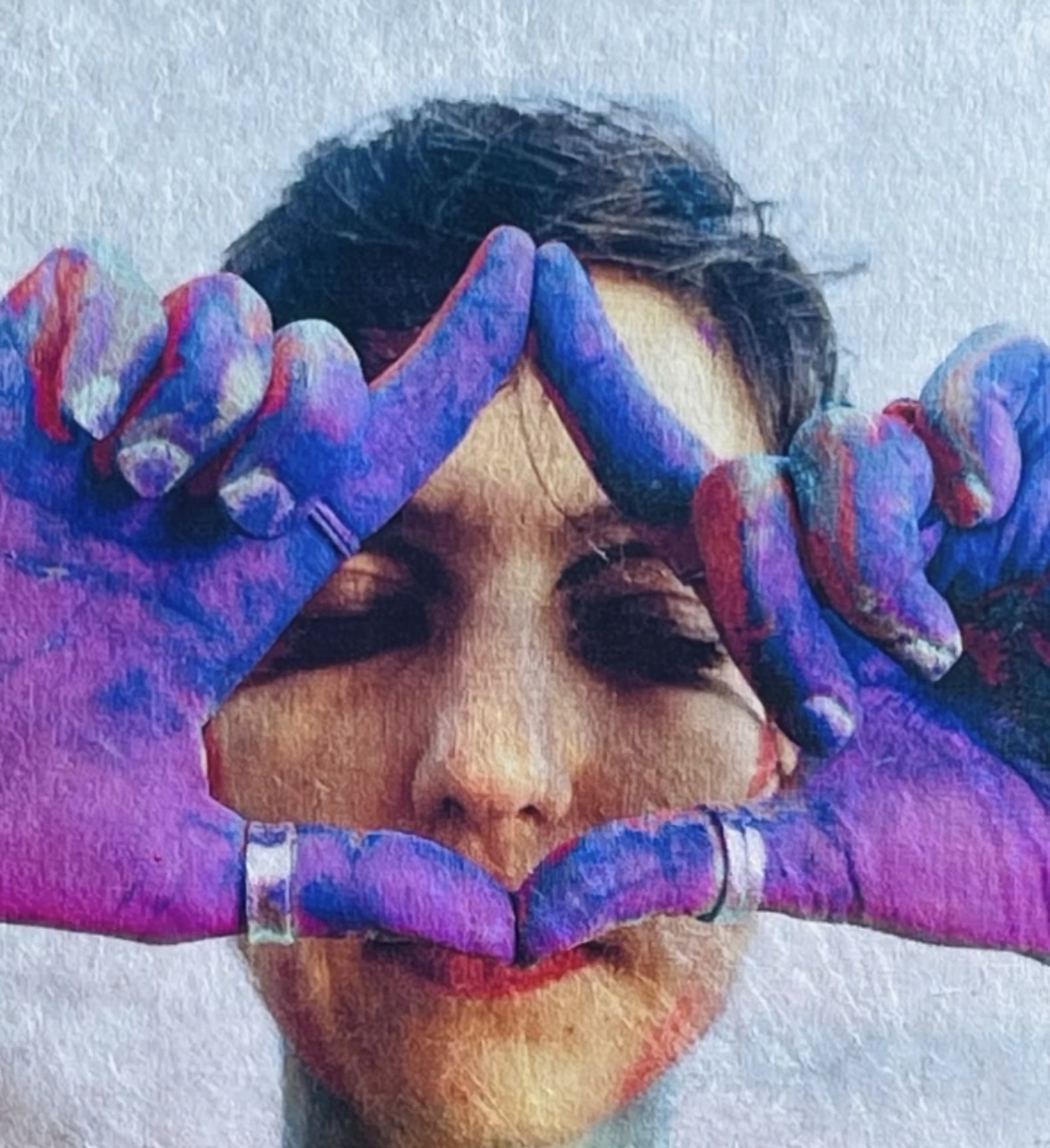 Person with hands covered in colorful paint forming a heart shape around their face, eyes closed, smiling.