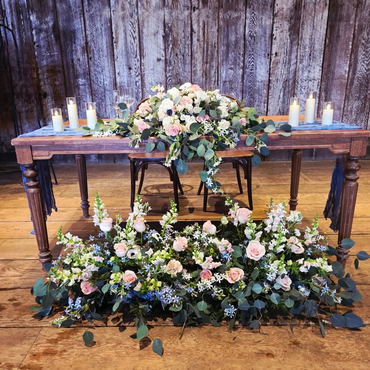 Floral arrangement with pink and white roses, greenery, and blue flowers on a wooden table with candles, against a wooden wall.