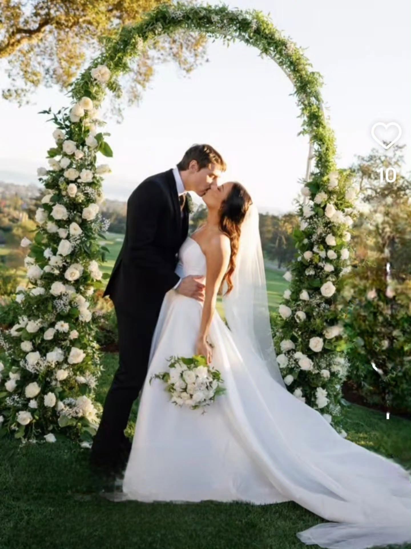 Bride and groom kiss under a floral arch outdoors, bride holding a bouquet, wedding dress flowing, trees in background.