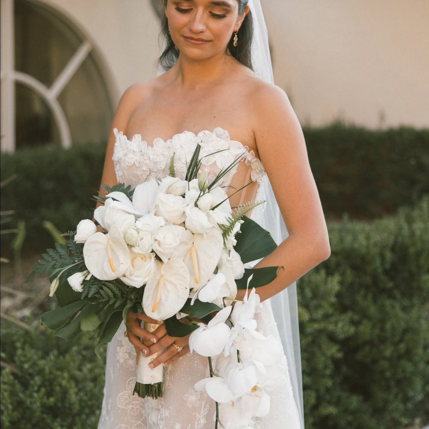 Bride holding a large bouquet of white flowers, wearing a strapless wedding dress, outdoors with greenery in background.