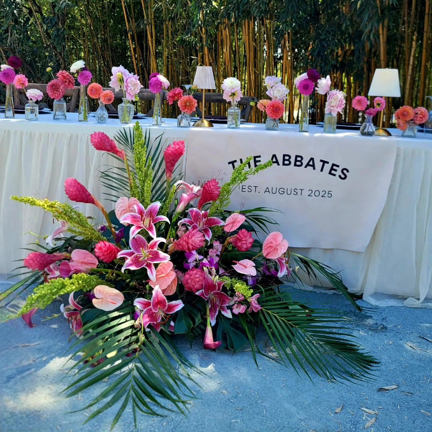 Flower arrangement with pink lilies, roses, and greenery in front of a table with pink and white flowers, and a sign reading 'THE ABBATES EST. AUGUST 2025'.