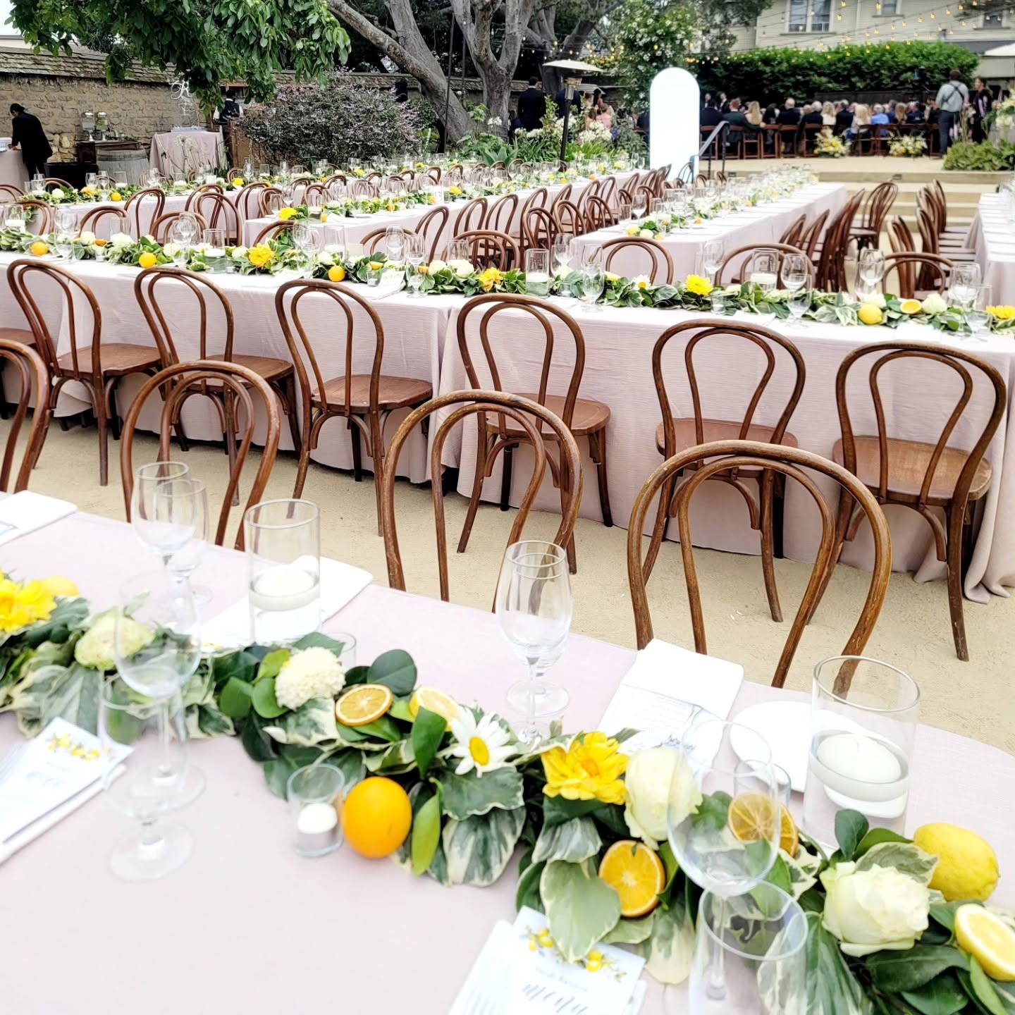 Long outdoor tables decorated with yellow and white flowers, lemons, and candles, set for a celebration or event.