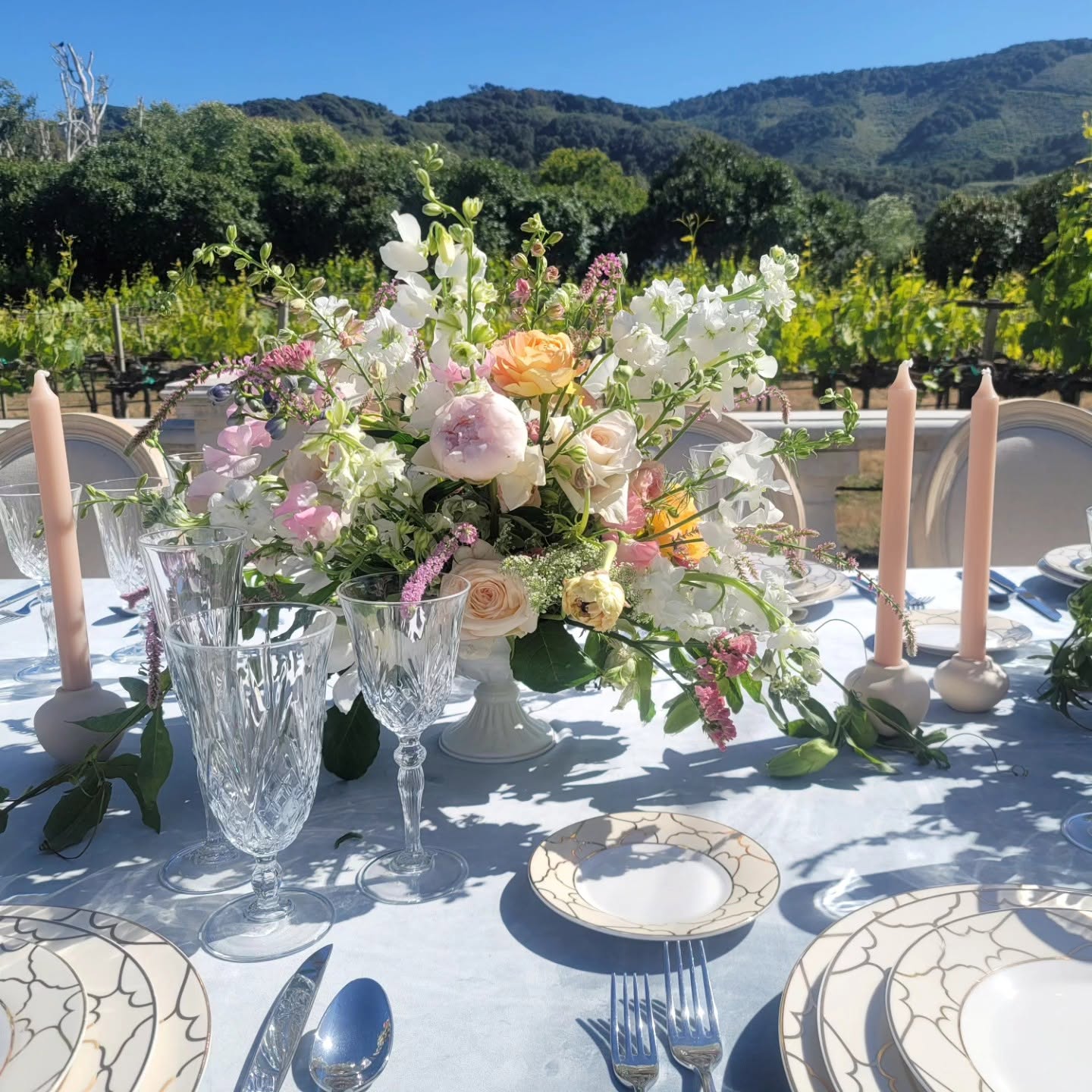 Table with a floral centerpiece, candles, plates, and cutlery outdoors with a scenic mountain view.