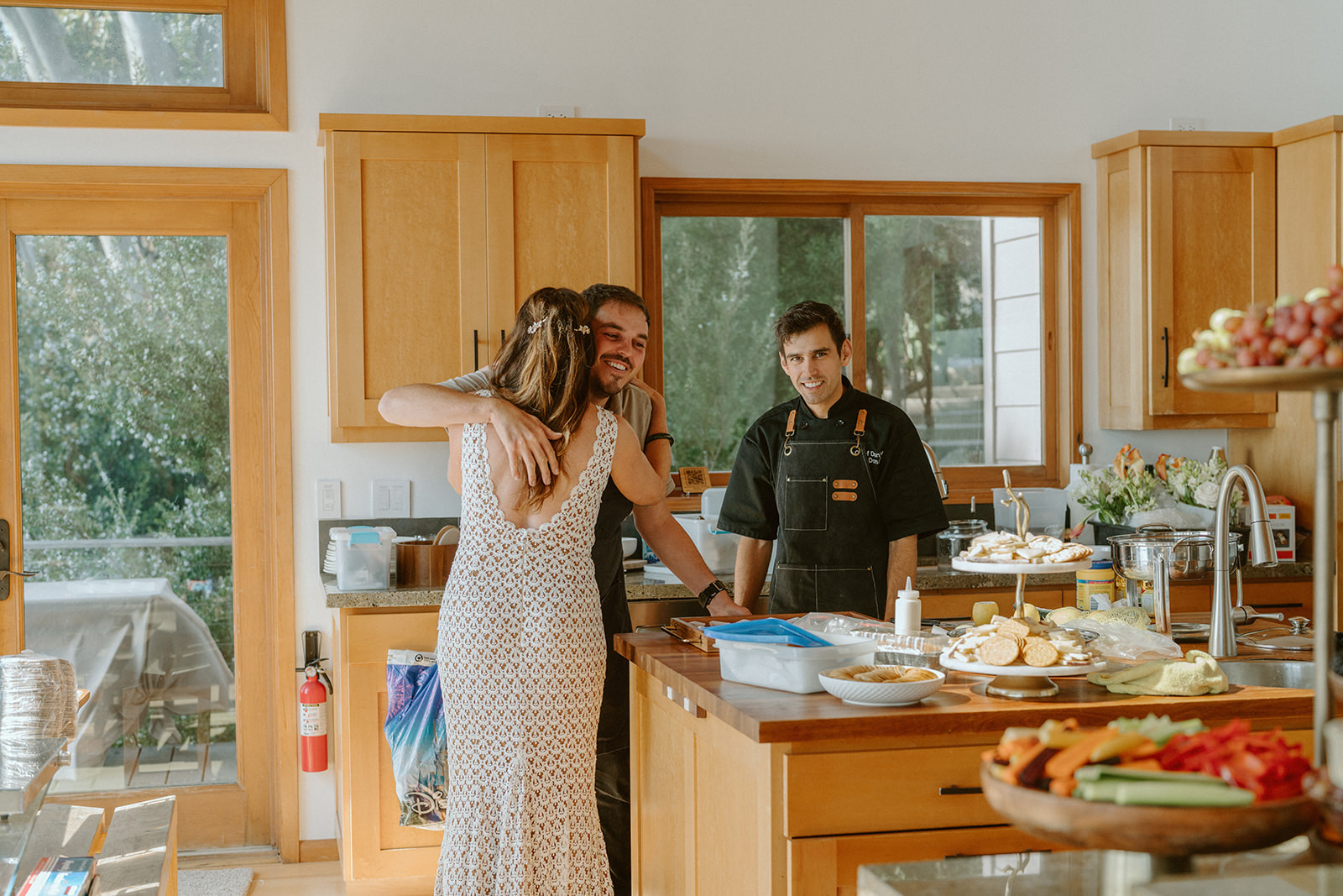 Three people in a kitchen, two women and one man, with wooden cabinets and a window showing outdoor greenery.