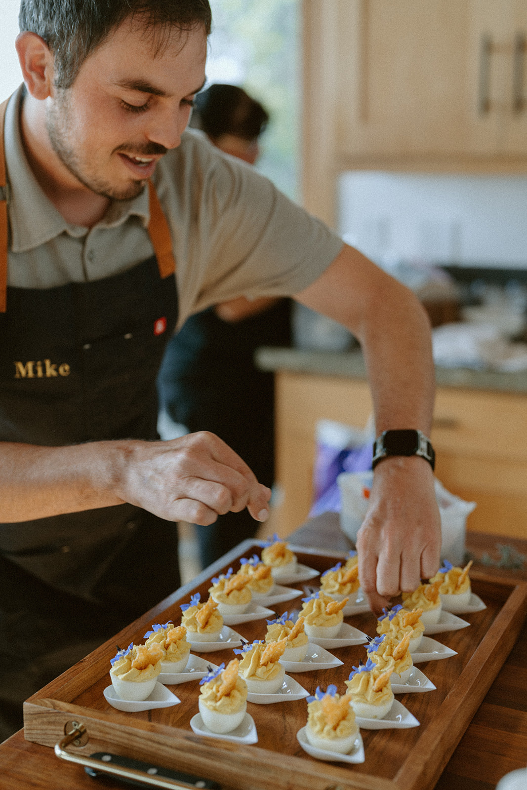 Man arranging small desserts on a tray in a kitchen, wearing an apron with the name 'Mike'.