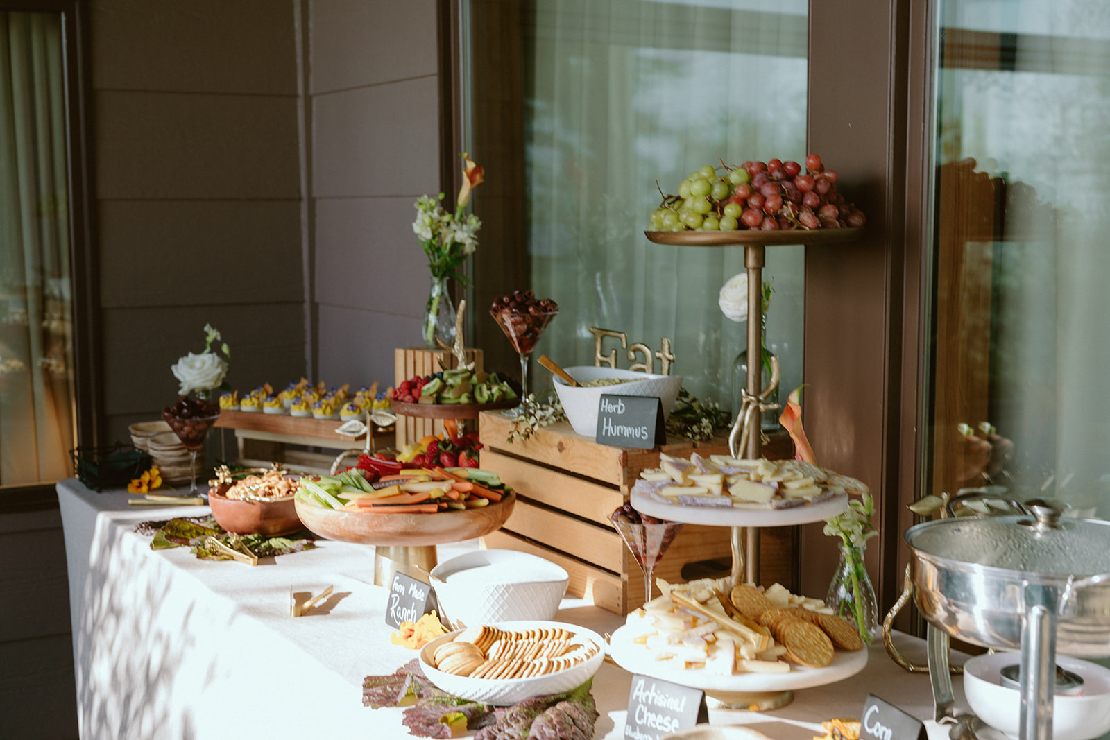 Table with various desserts and fruits, decorated with flowers and a wooden crate, set near a window.