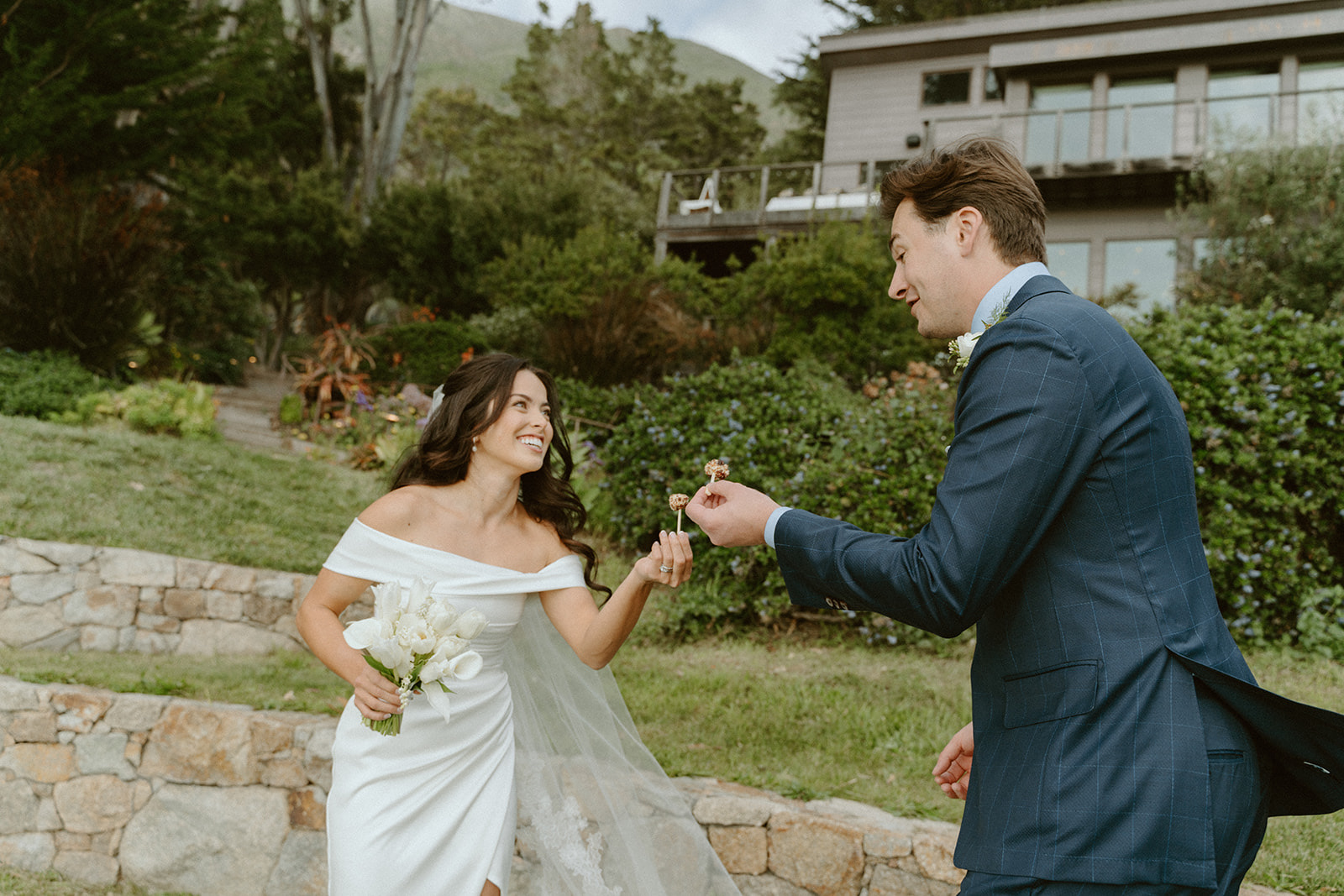 A bride in a white wedding dress and a groom in a blue suit exchange rings outdoors, holding hands, with greenery and a building in background.