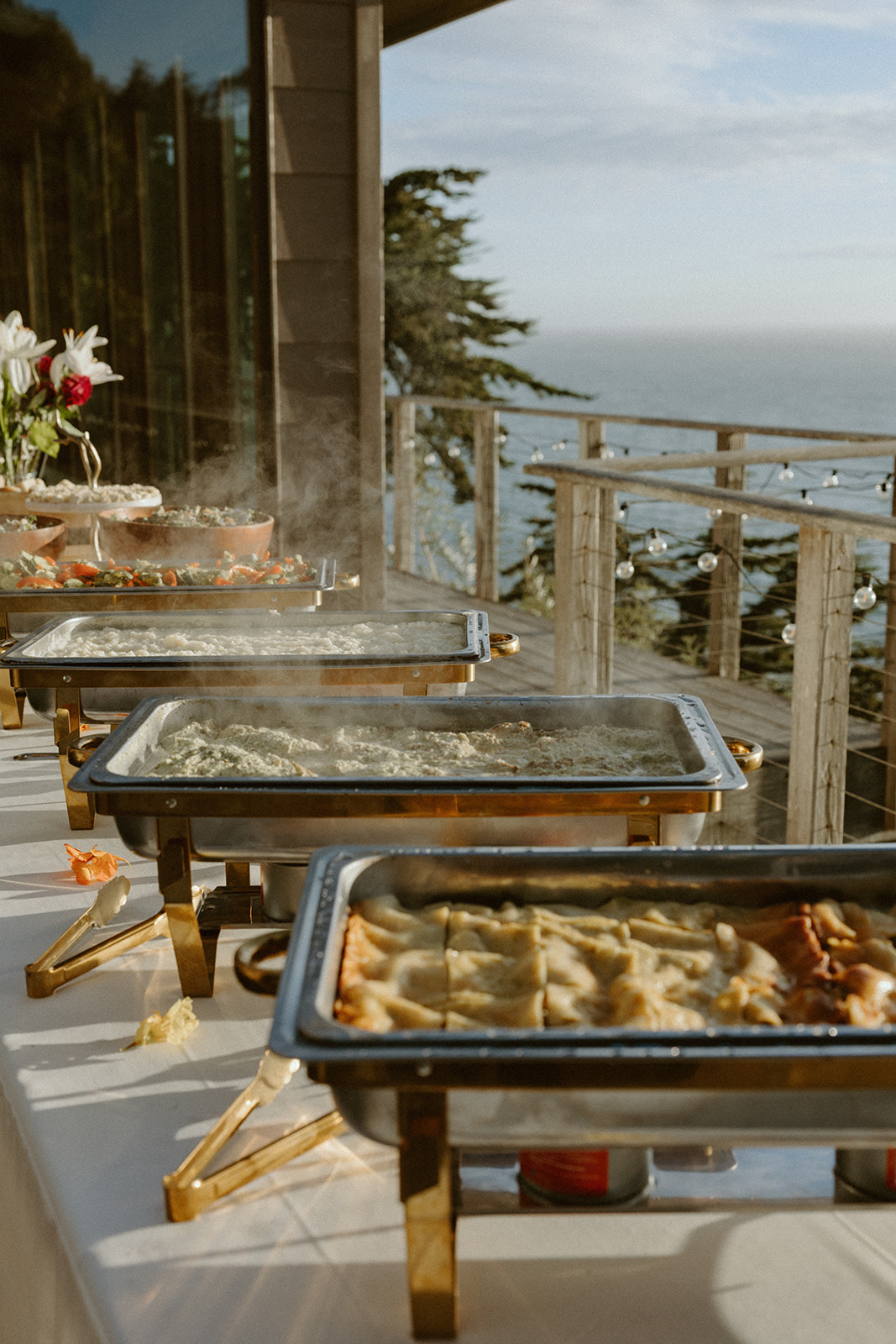 Buffet setup with chafing dishes on a balcony overlooking the ocean, with a railing and trees in the background.