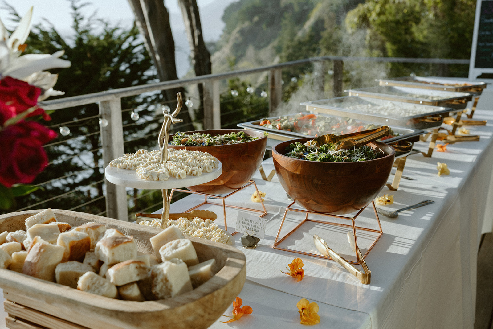 Buffet table with bowls of salad and dishes, outdoor setting with trees and railing in background.