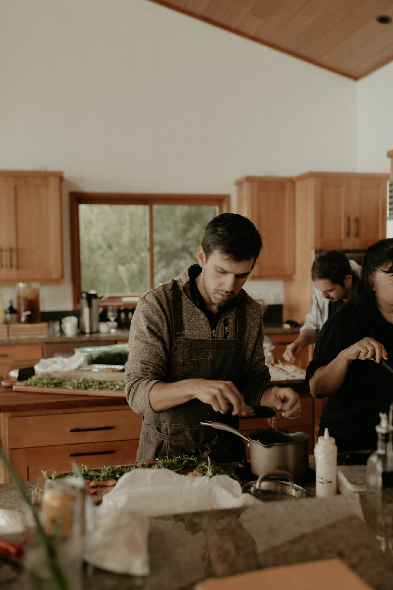 Man cooking in a kitchen with wooden cabinets and a window, surrounded by ingredients and utensils.
