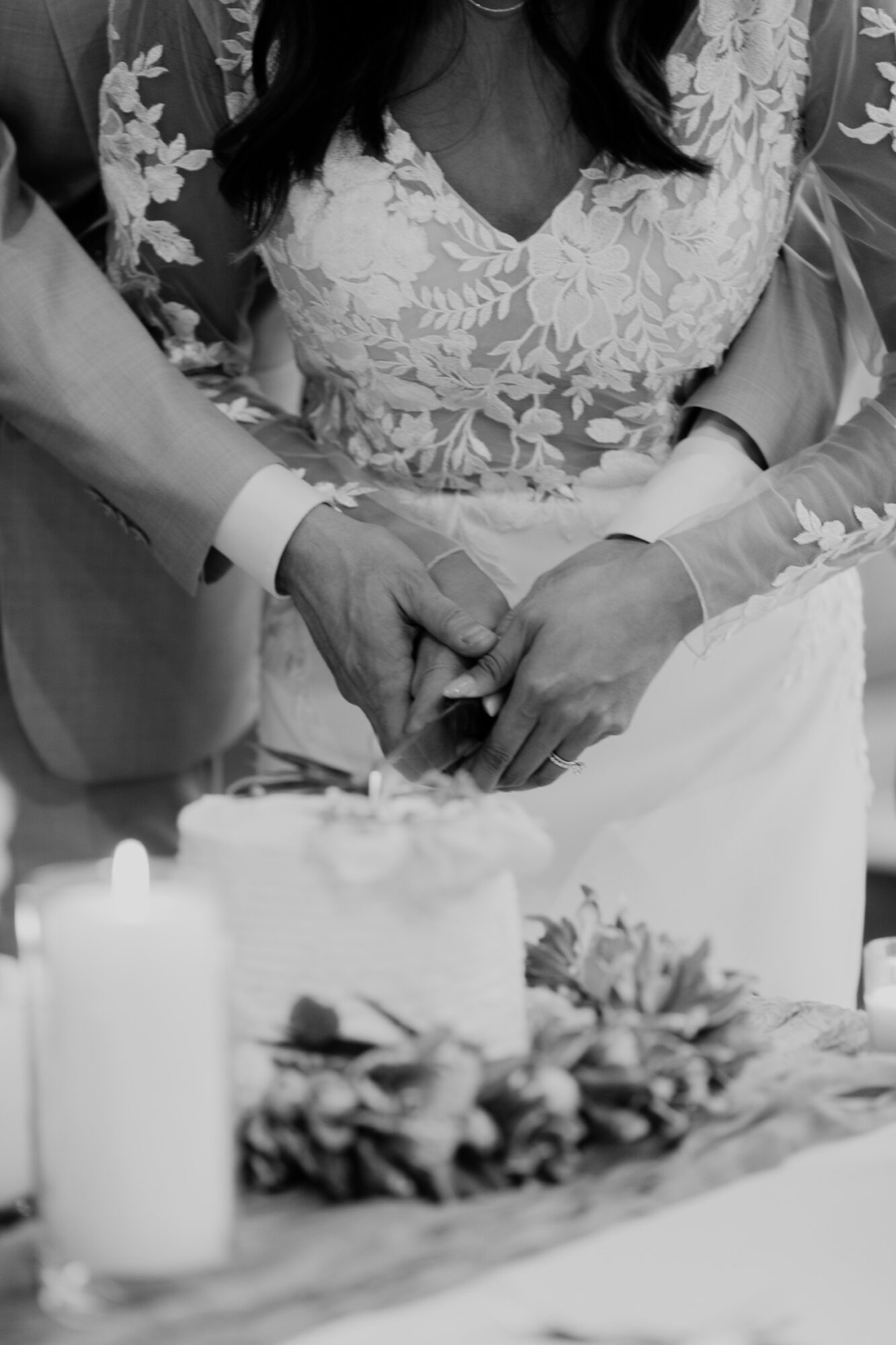 Two people hold hands over a wedding cake, with floral decorations and candles in the foreground.