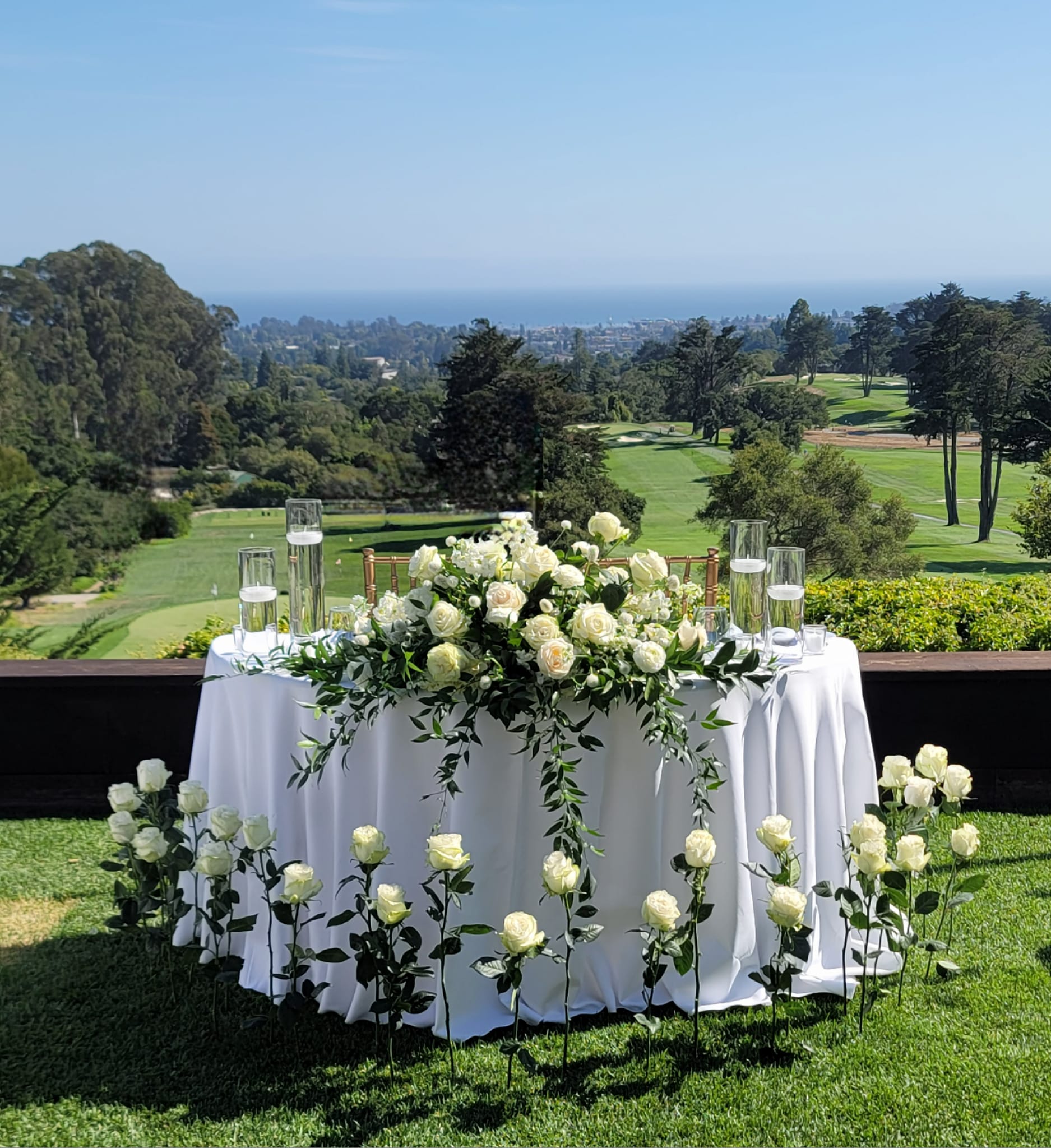 A round table with a white tablecloth and floral centerpiece, set outdoors with a golf course and trees in the background.