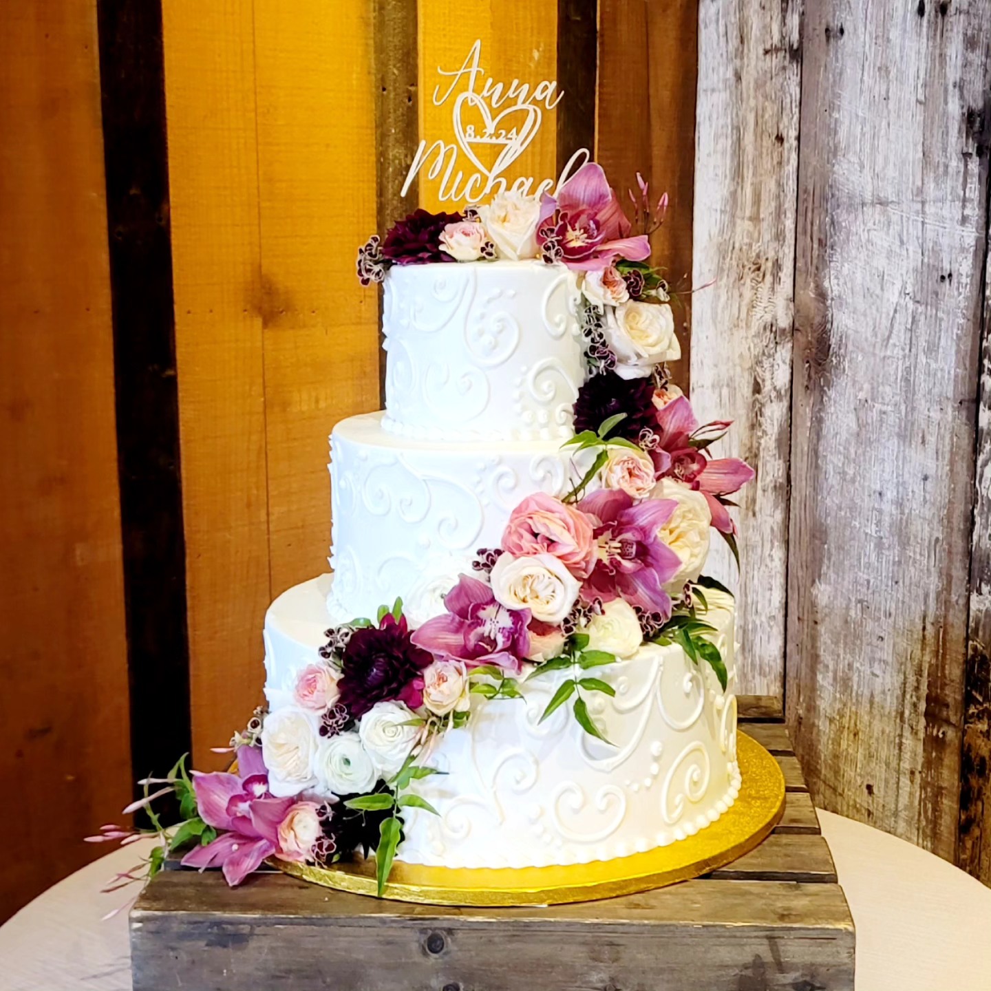 Three-tier white wedding cake decorated with pink and white flowers, on a wooden table with rustic background.