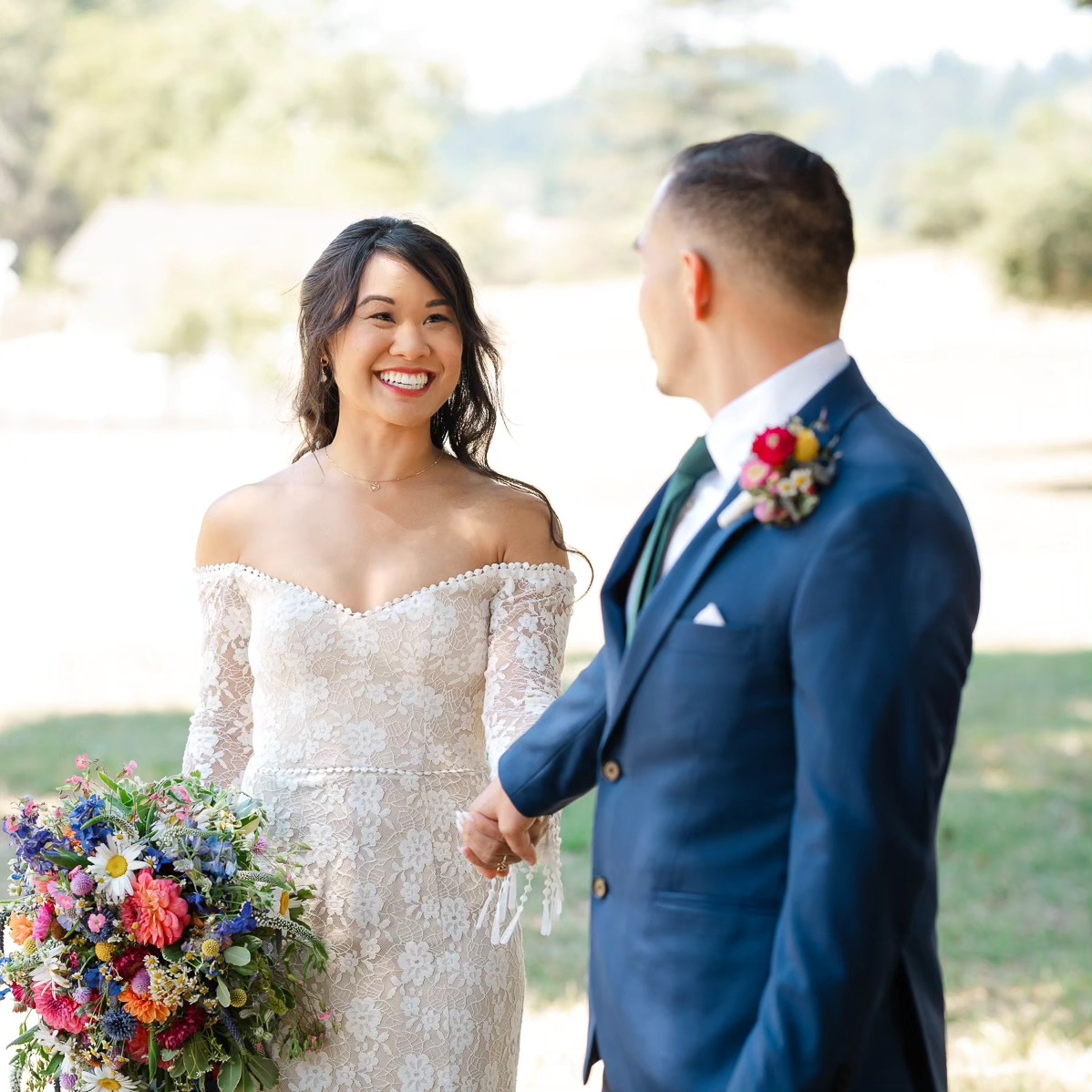 A bride and groom holding hands outdoors, smiling at each other, with a scenic background. The bride wears a lace off-shoulder dress, holding a colorful bouquet. The groom wears a blue suit with a boutonniere.